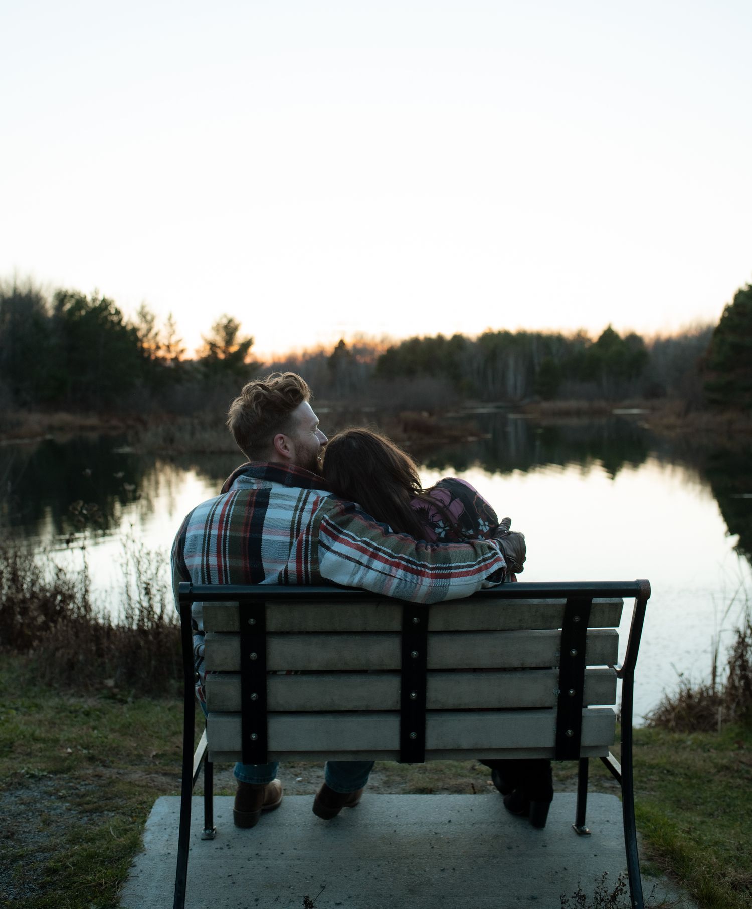 Two people in matching plaid shirts sit together on a bench by a serene lake at sunset.