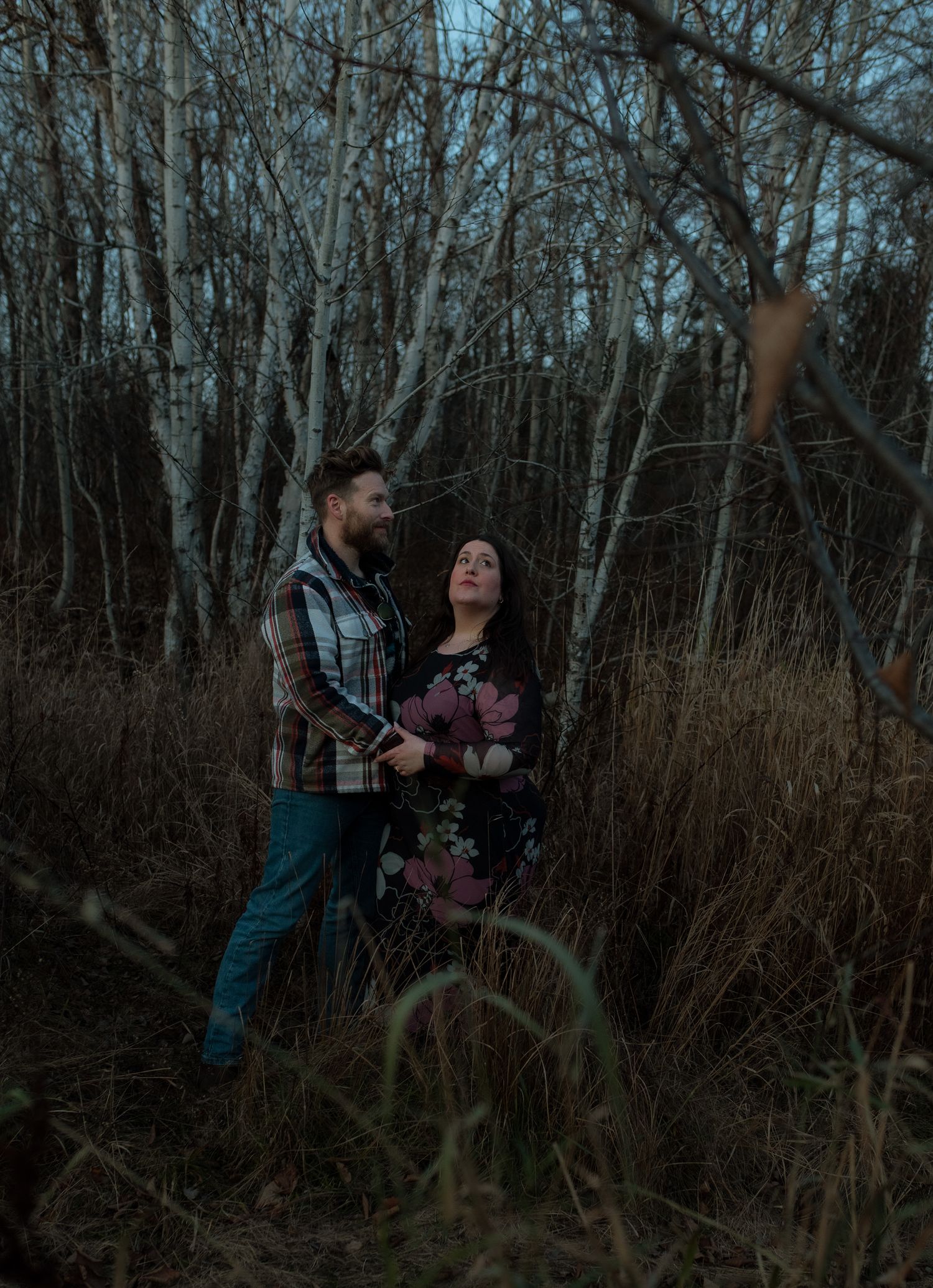 A couple shares a romantic moment among bare winter trees and tall grass at dusk.
