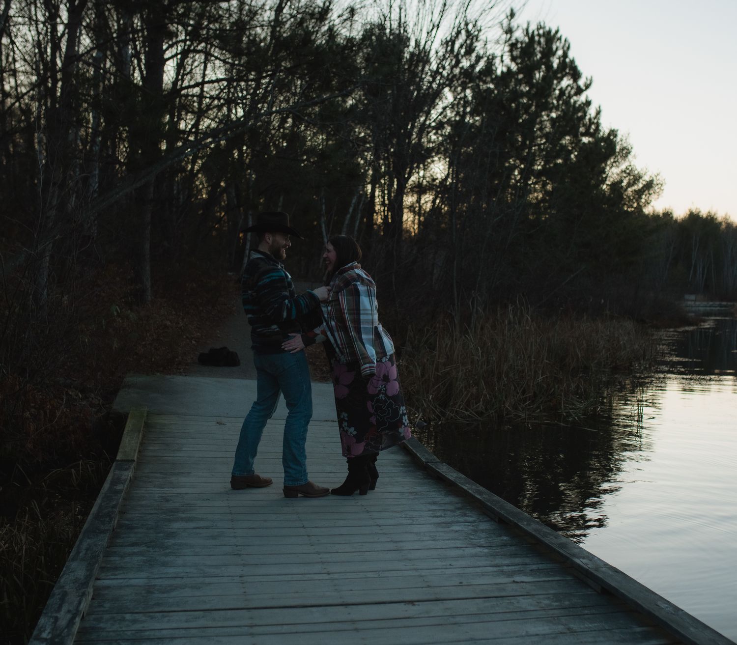 People gather on a wooden boardwalk at sunset overlooking a serene lake surrounded by trees.