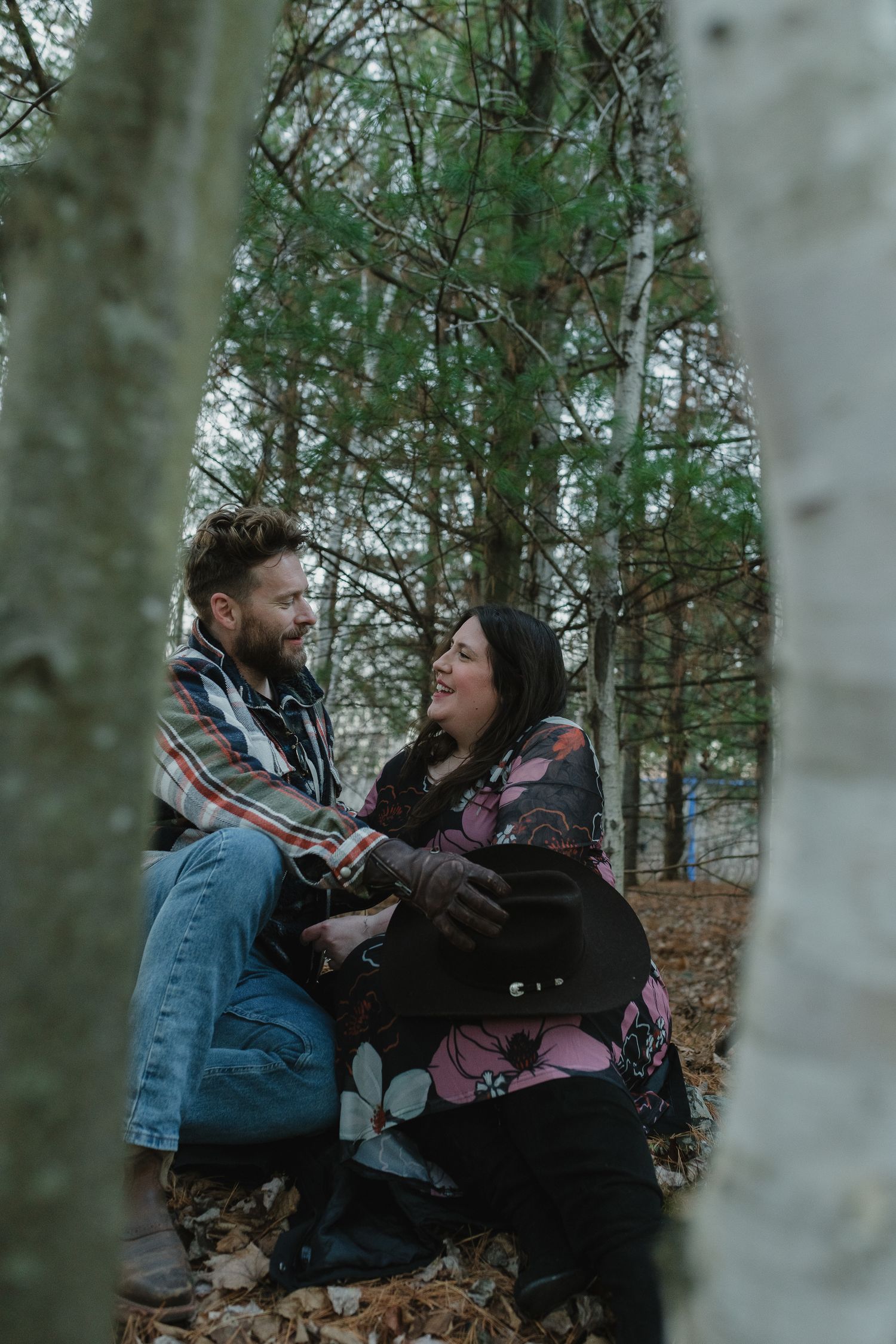 A couple sits together among tall trees in a forest during an intimate outdoor photoshoot.