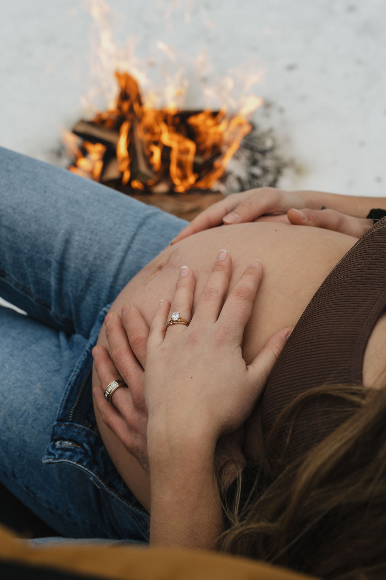 Hands rest on a pregnant belly near a glowing campfire in the snow.