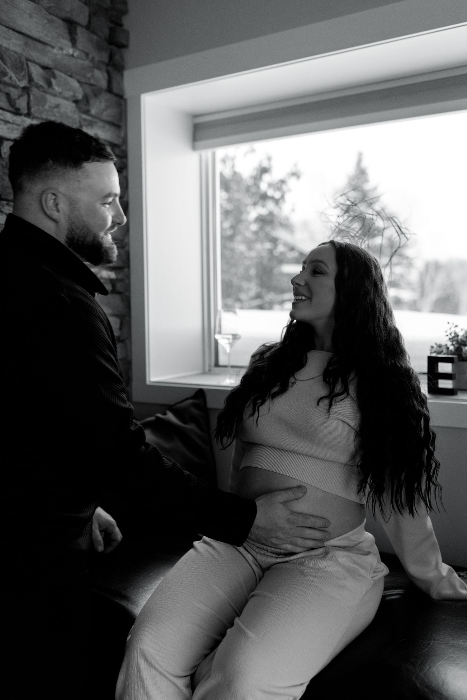 Black and white maternity photo of couple sitting by a window, woman cradling her baby bump.