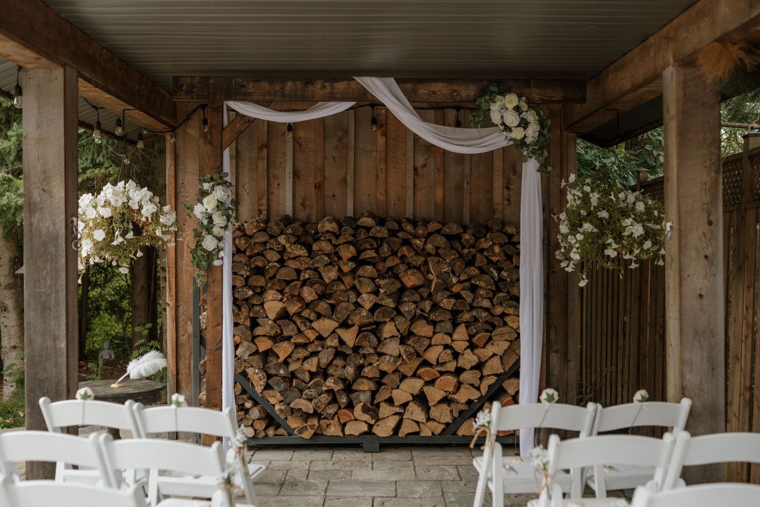 Rustic outdoor wedding ceremony setup with white chairs, draped fabric, floral arrangements, and a stacked firewood backdrop.