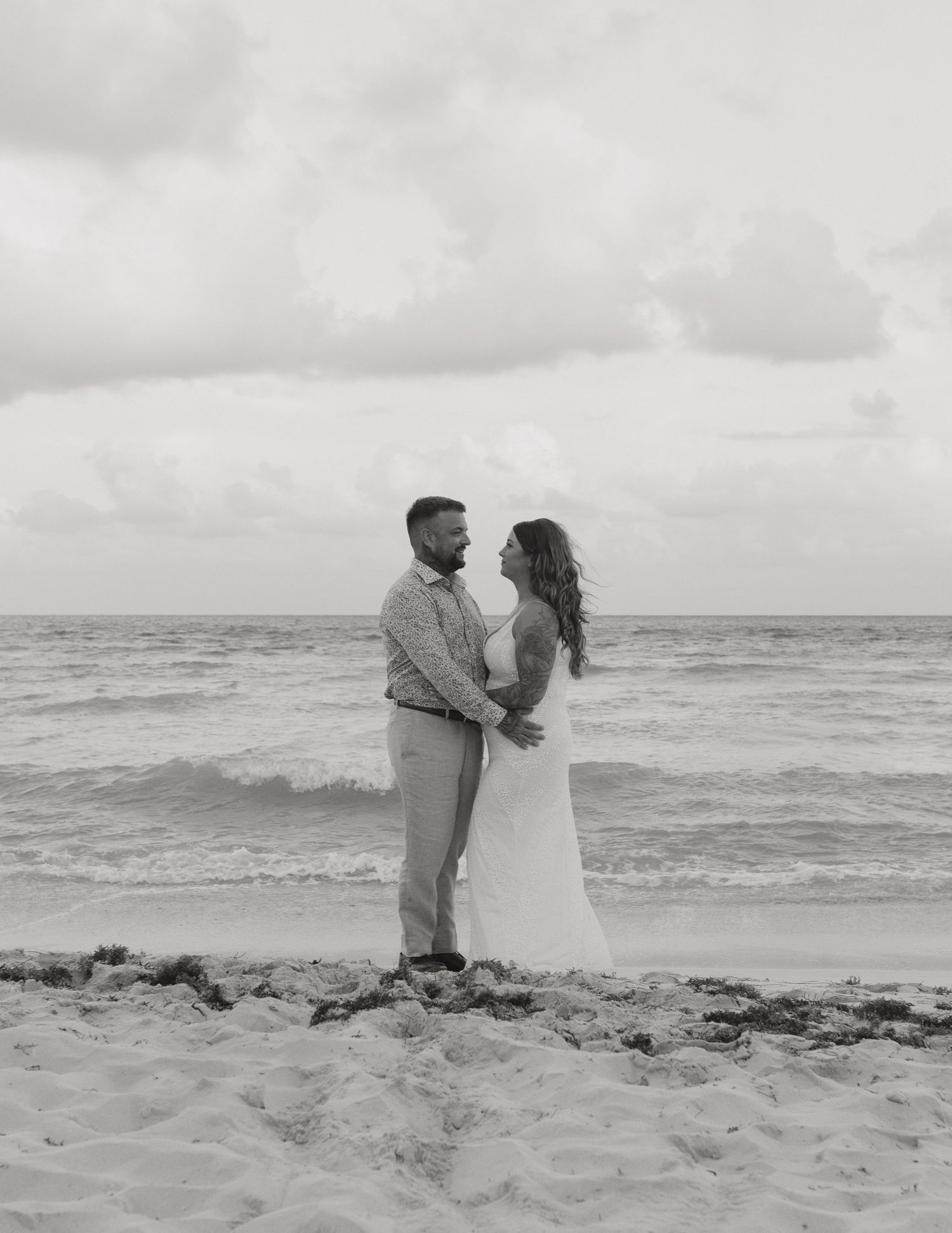 Black and white beach wedding photo of couple embracing by ocean waves, bride in white gown, groom in casual attire.