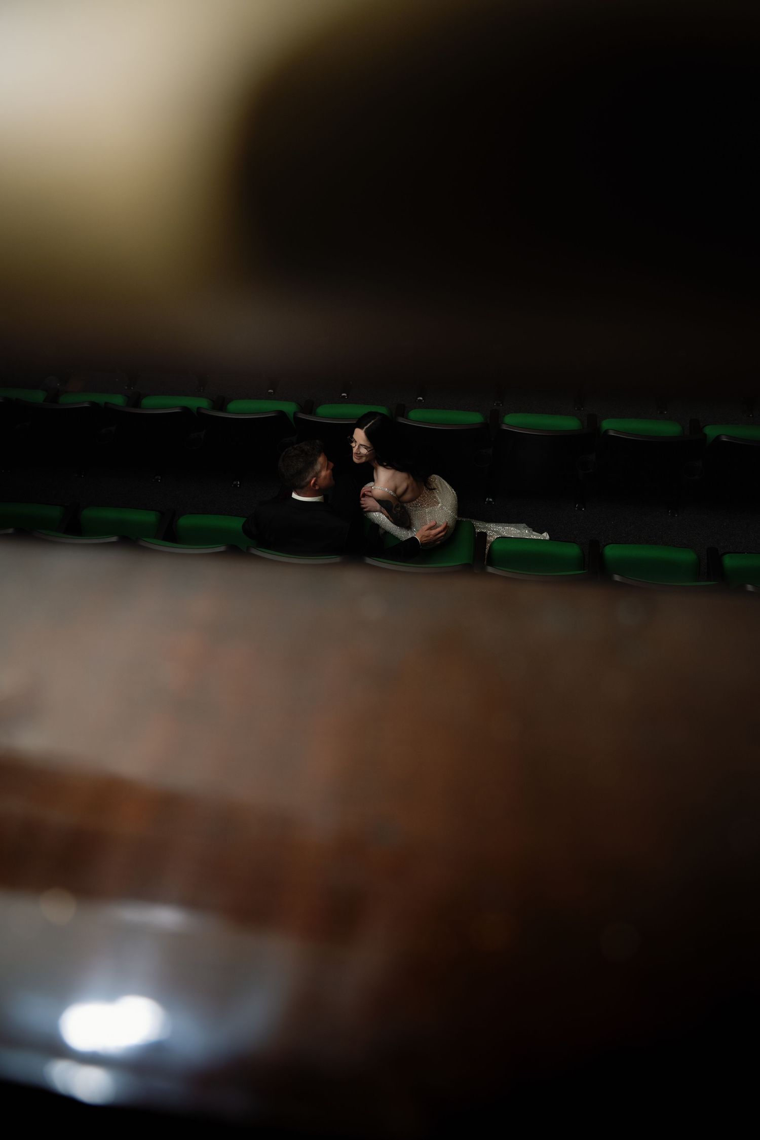 Couple sharing an intimate moment in dimly lit theater seats, viewed from above between rows.