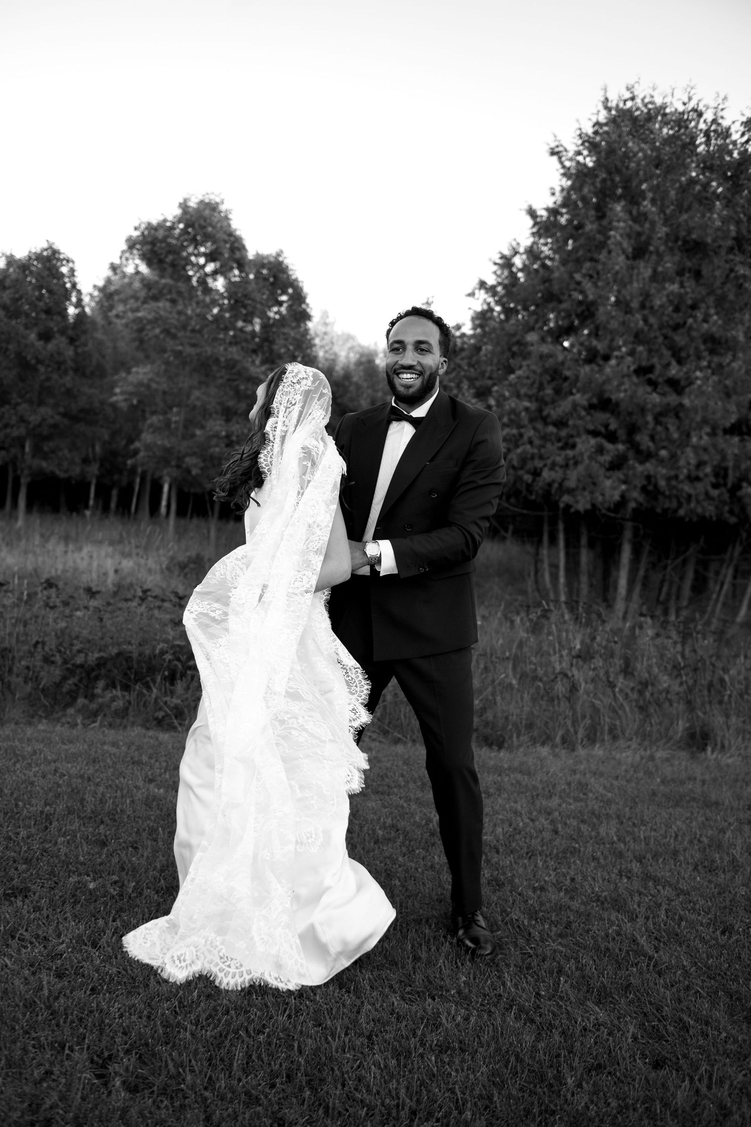 Black and white photo of smiling groom in tuxedo dancing with bride in lace wedding dress in an outdoor field.