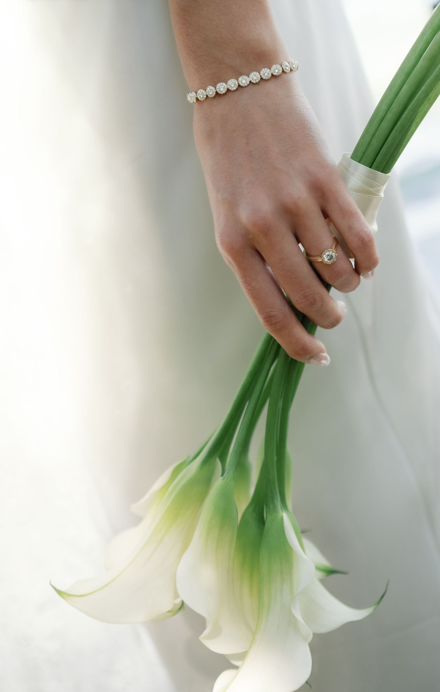 Bride holding elegant white calla lily bouquet, wearing pearl bracelet and diamond ring, in soft natural light.