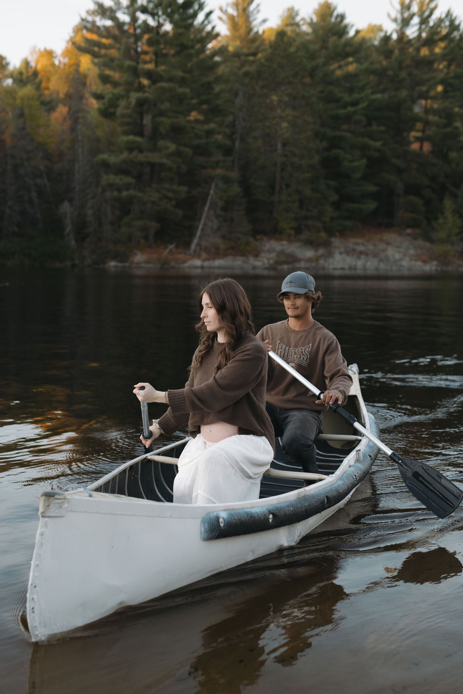 Two people paddle together in a white canoe on a calm lake surrounded by pine trees.