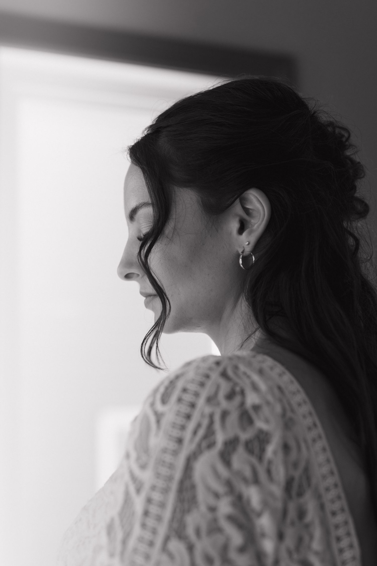 Black and white profile portrait of a bride with dark hair in lace dress, looking down near a bright window.