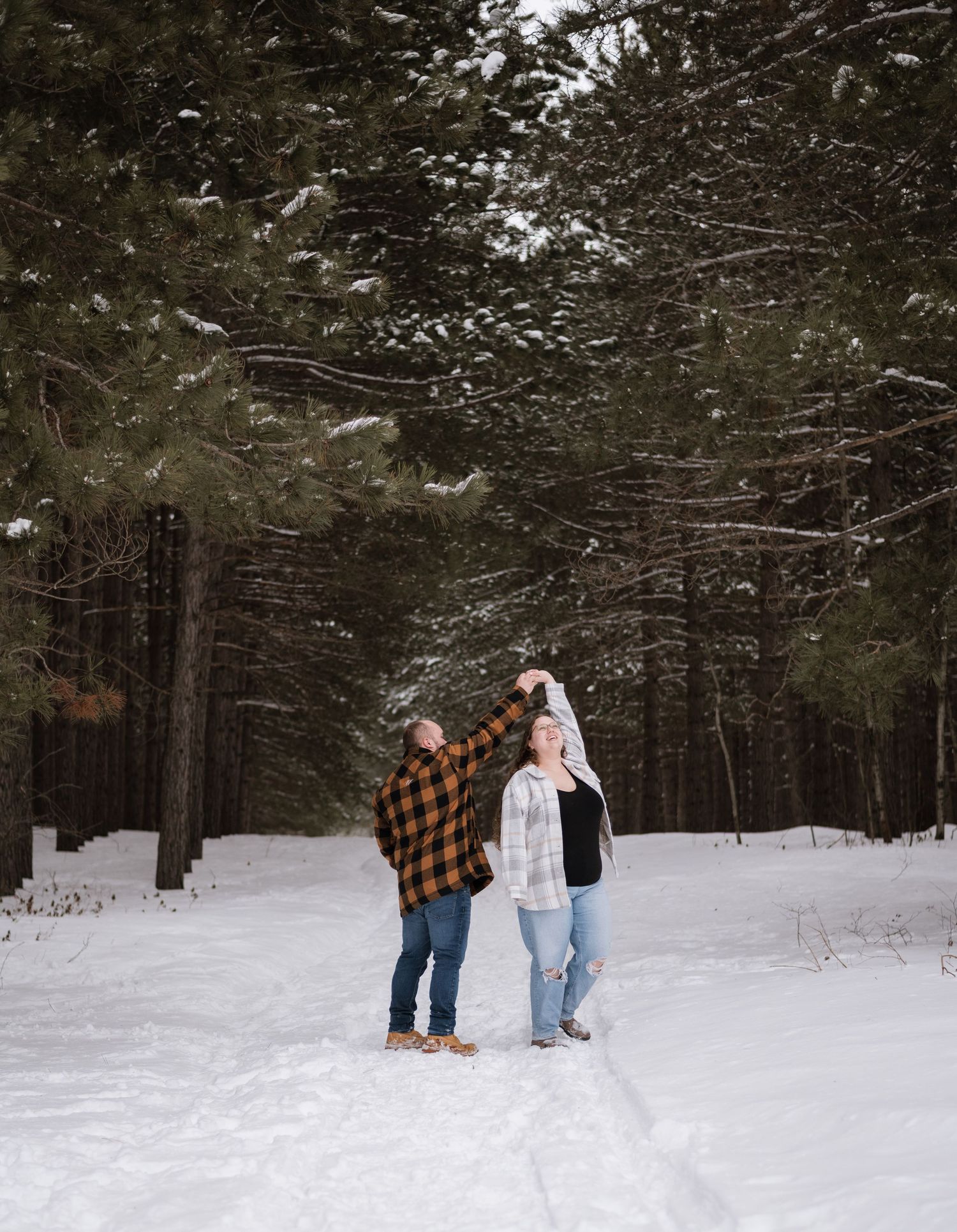 Couple dancing in snowy forest path surrounded by tall pine trees in winter, wearing plaid flannel shirts and jeans.