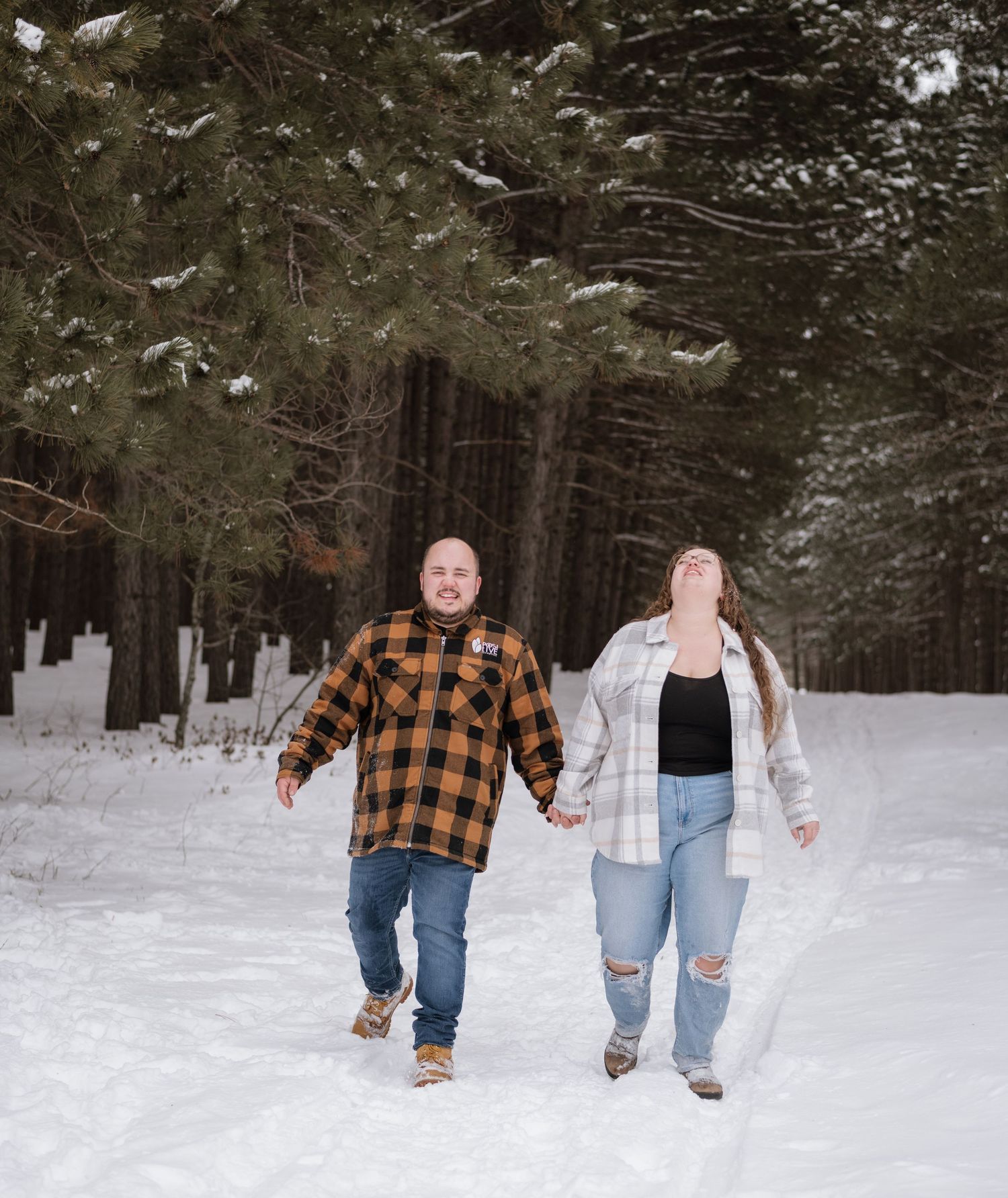 Couple joyfully running hand in hand down a snowy tree-lined path in plaid flannel shirts.