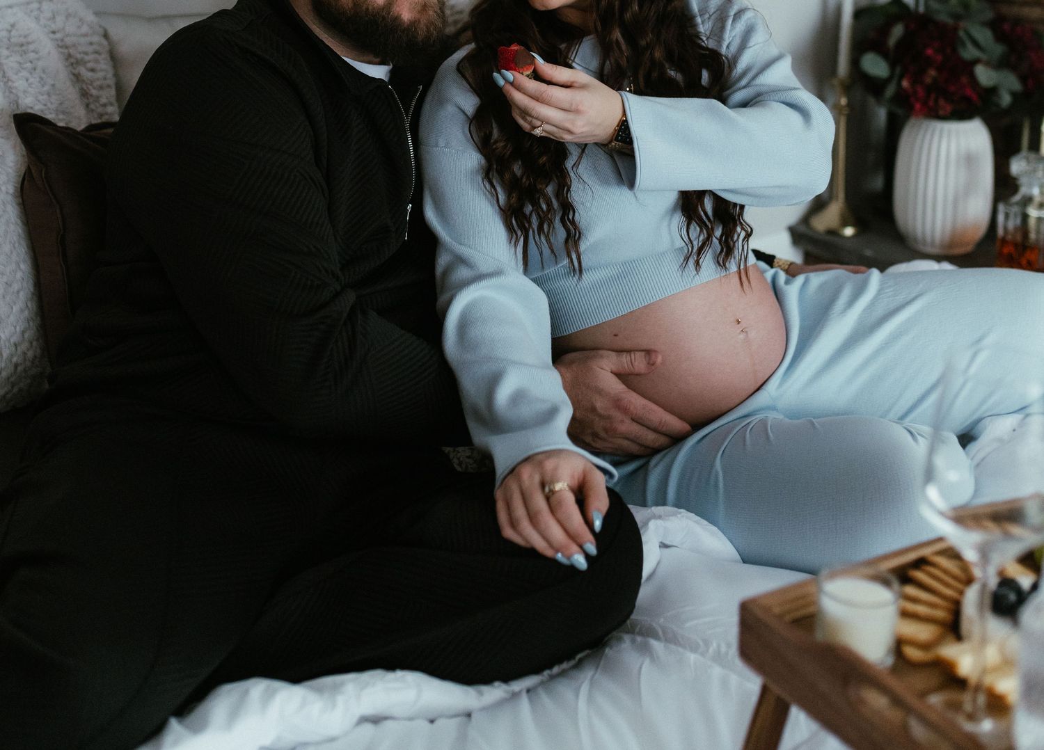 Pregnant couple sitting together on bed, man touching baby bump while woman eats strawberry.