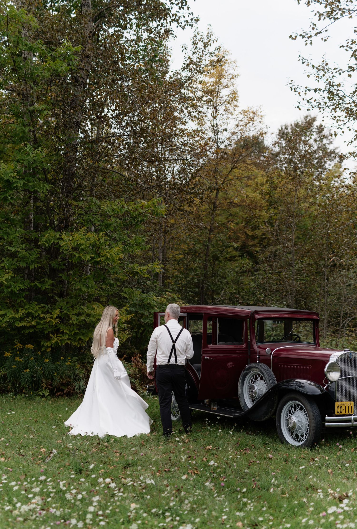 Bride and groom stand beside a vintage burgundy classic car surrounded by autumn trees at an outdoor wedding.
