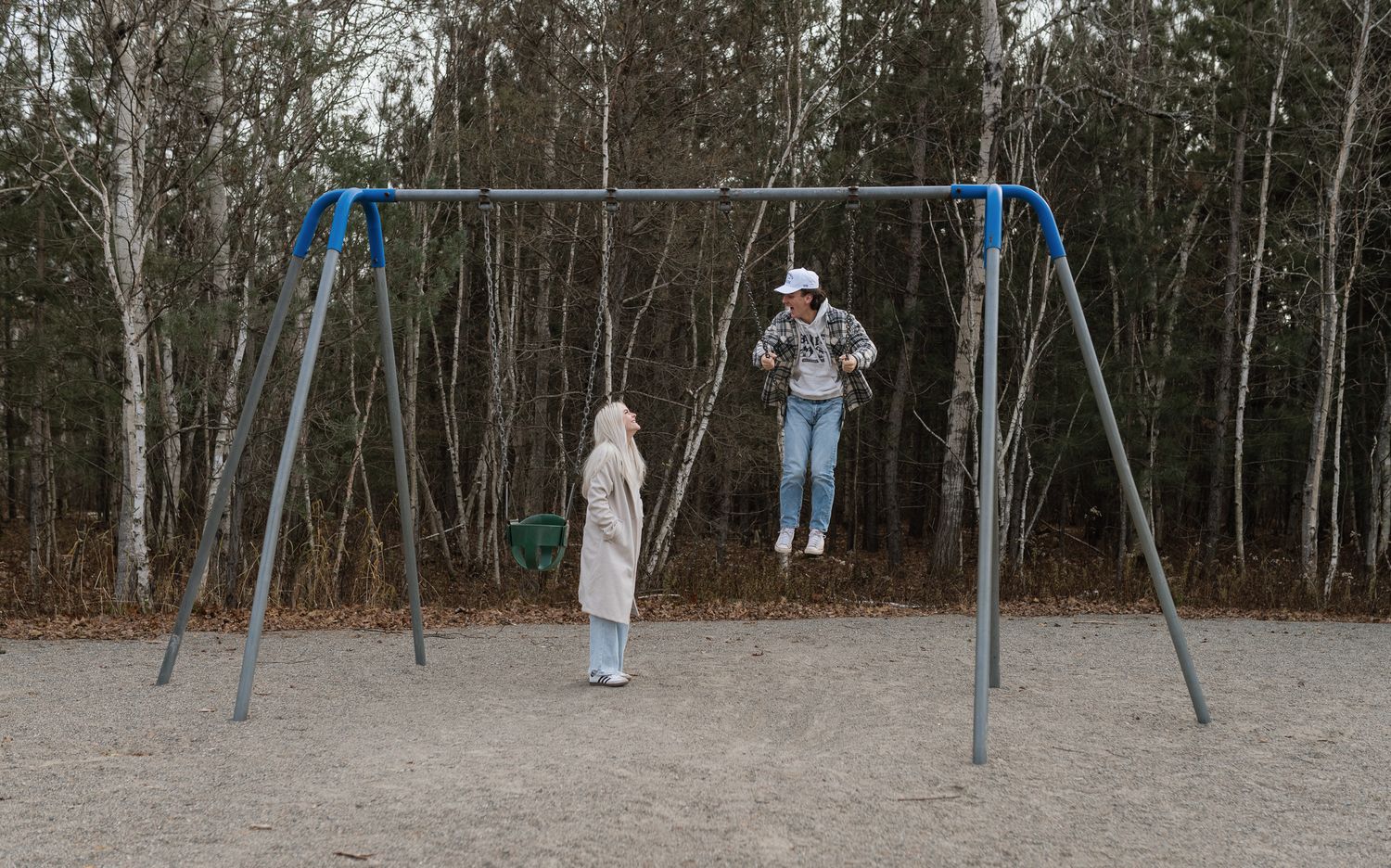 Child watches another child swinging on a blue metal swing set in a sandy playground surrounded by bare trees.