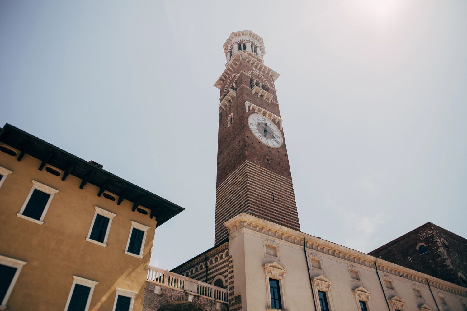 Tall medieval brick clock tower rising above historic Italian buildings against a bright sky in Verona.