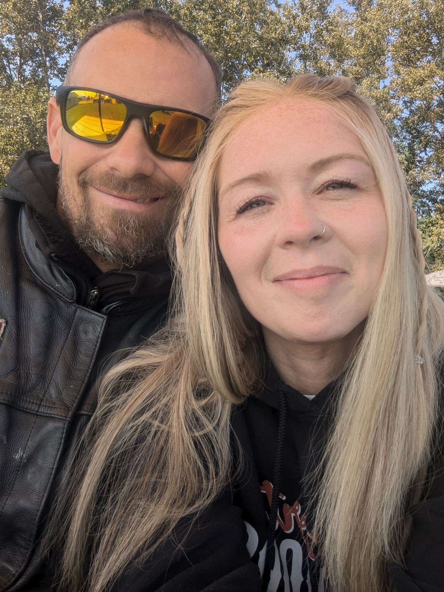 A smiling couple poses for a selfie outdoors, man with beard and yellow sunglasses, woman with long blonde hair.