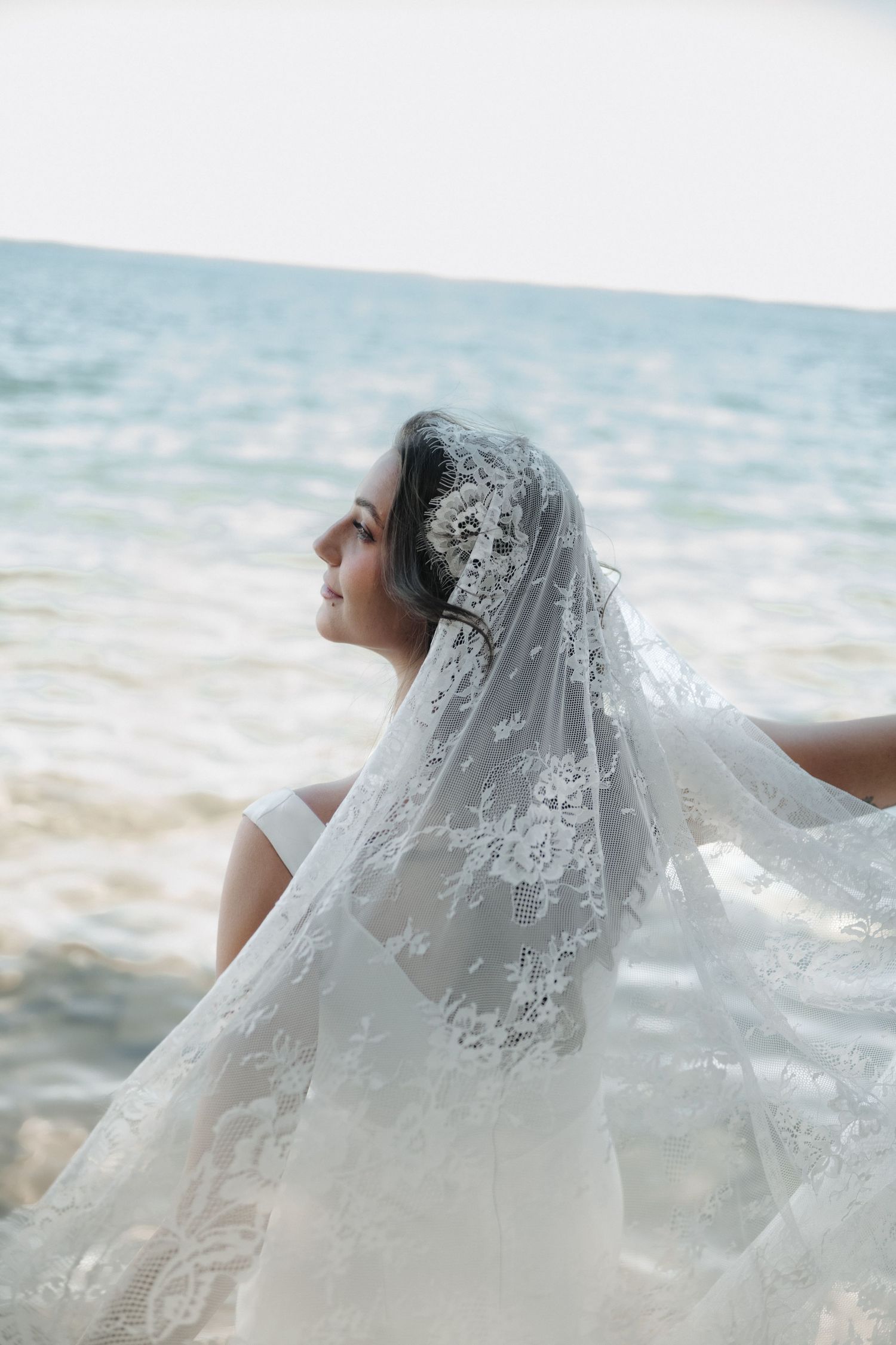 Bride in lace veil and gown gazing at ocean on beach, elegant bridal portrait with floral lace details.