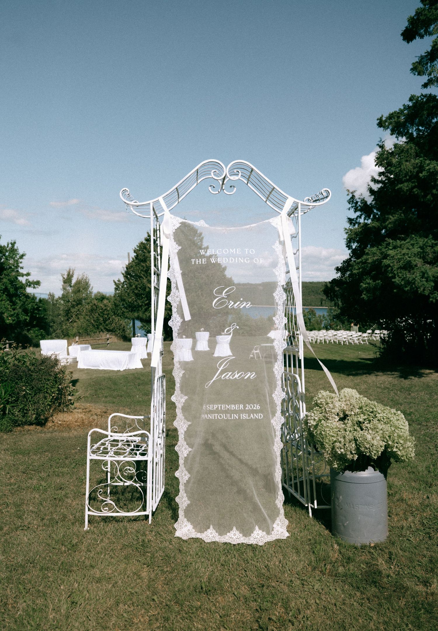 White ornate wedding arch with lace veil displaying Erin and Jason wedding welcome sign on sunny day.