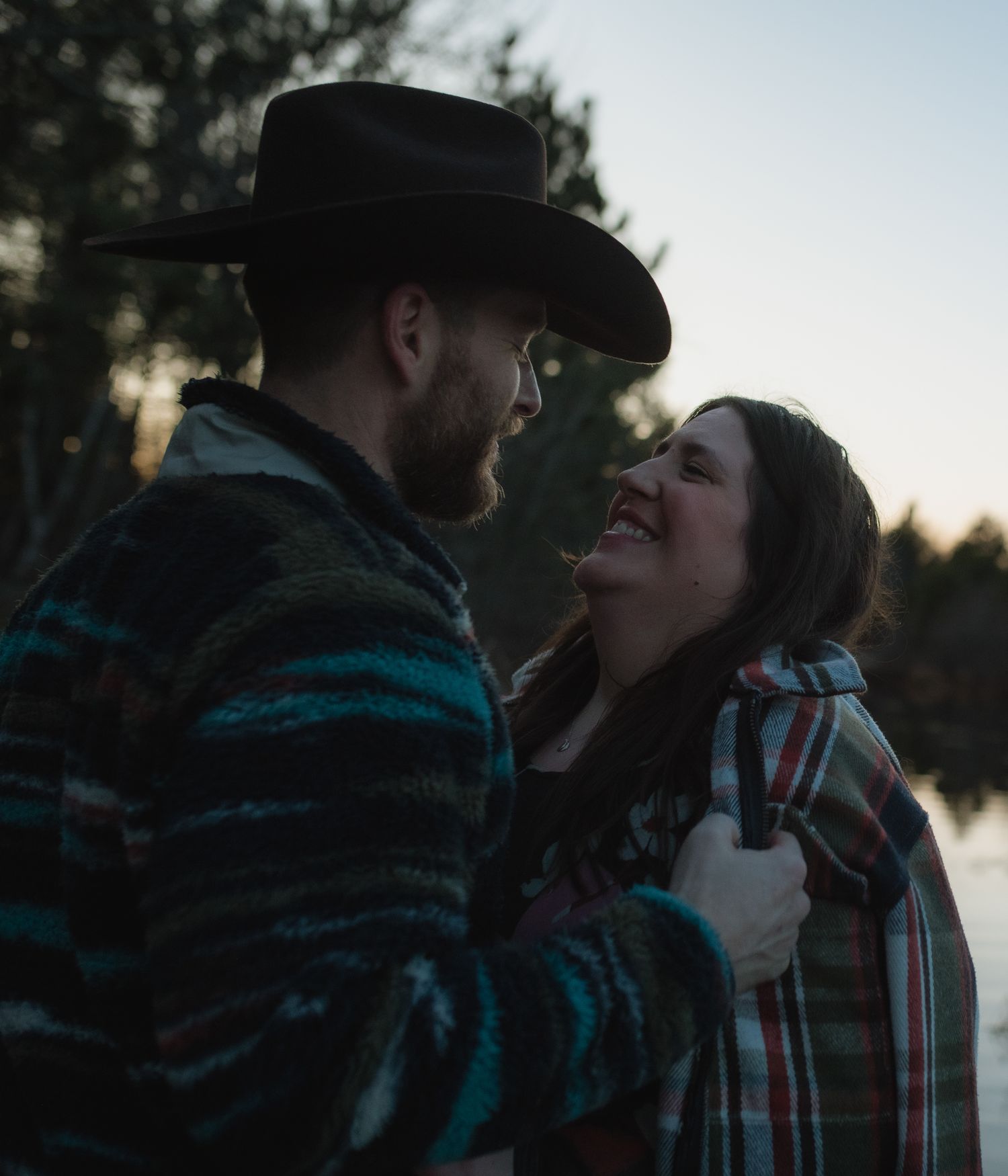A couple in winter clothing sharing an intimate moment outdoors at dusk, with a lake and trees in the background.