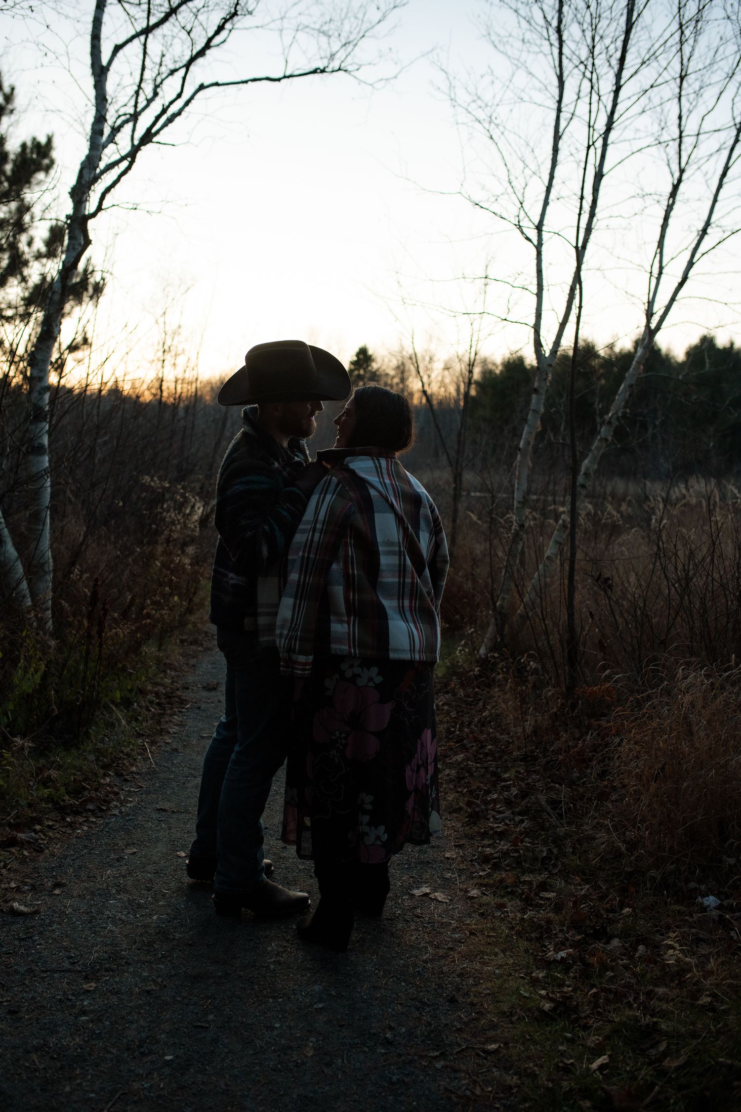 Two people in plaid jackets walk along a wooded trail at dusk during autumn.