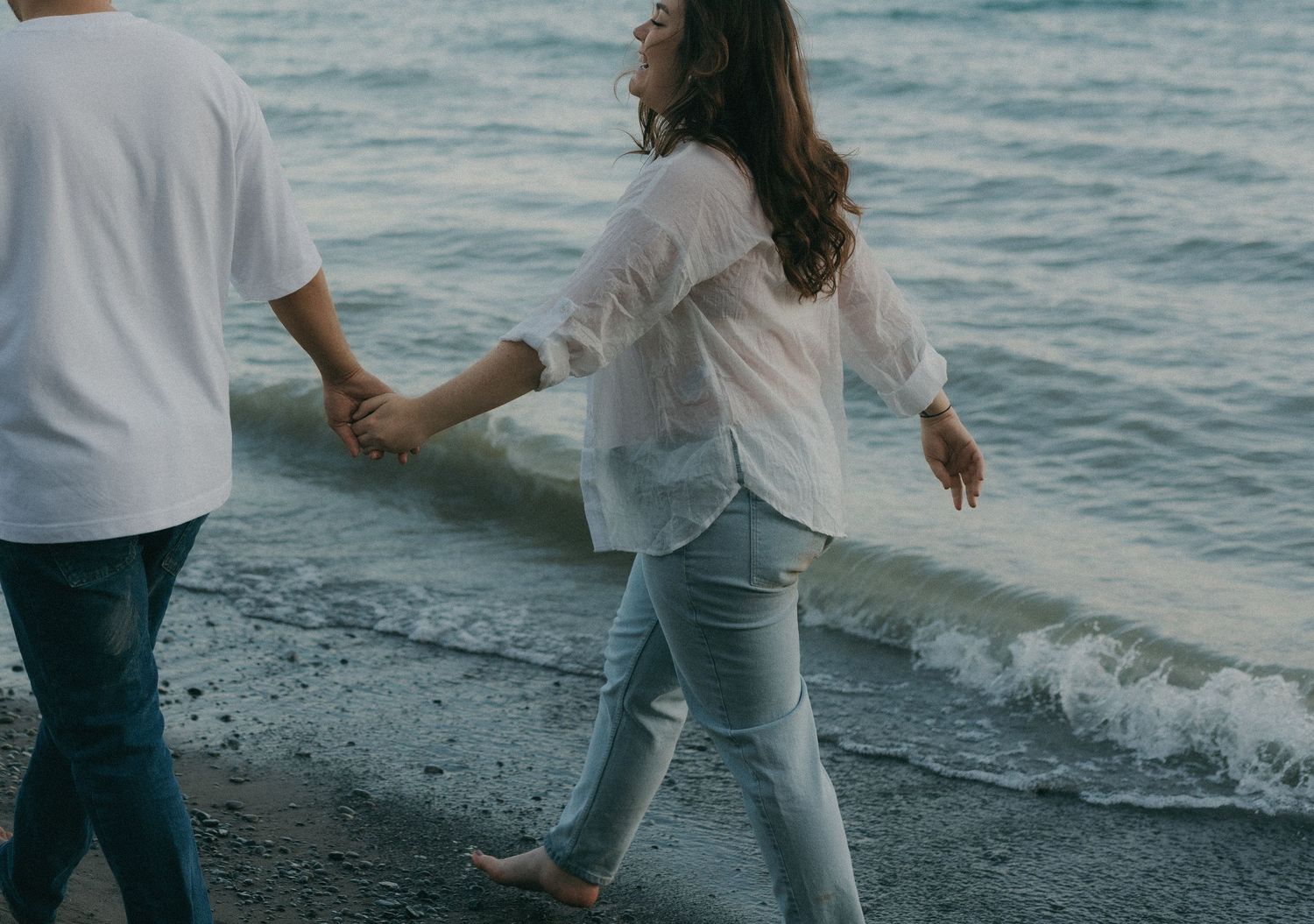 Couple walking along a rocky beach at dusk with waves crashing nearby.