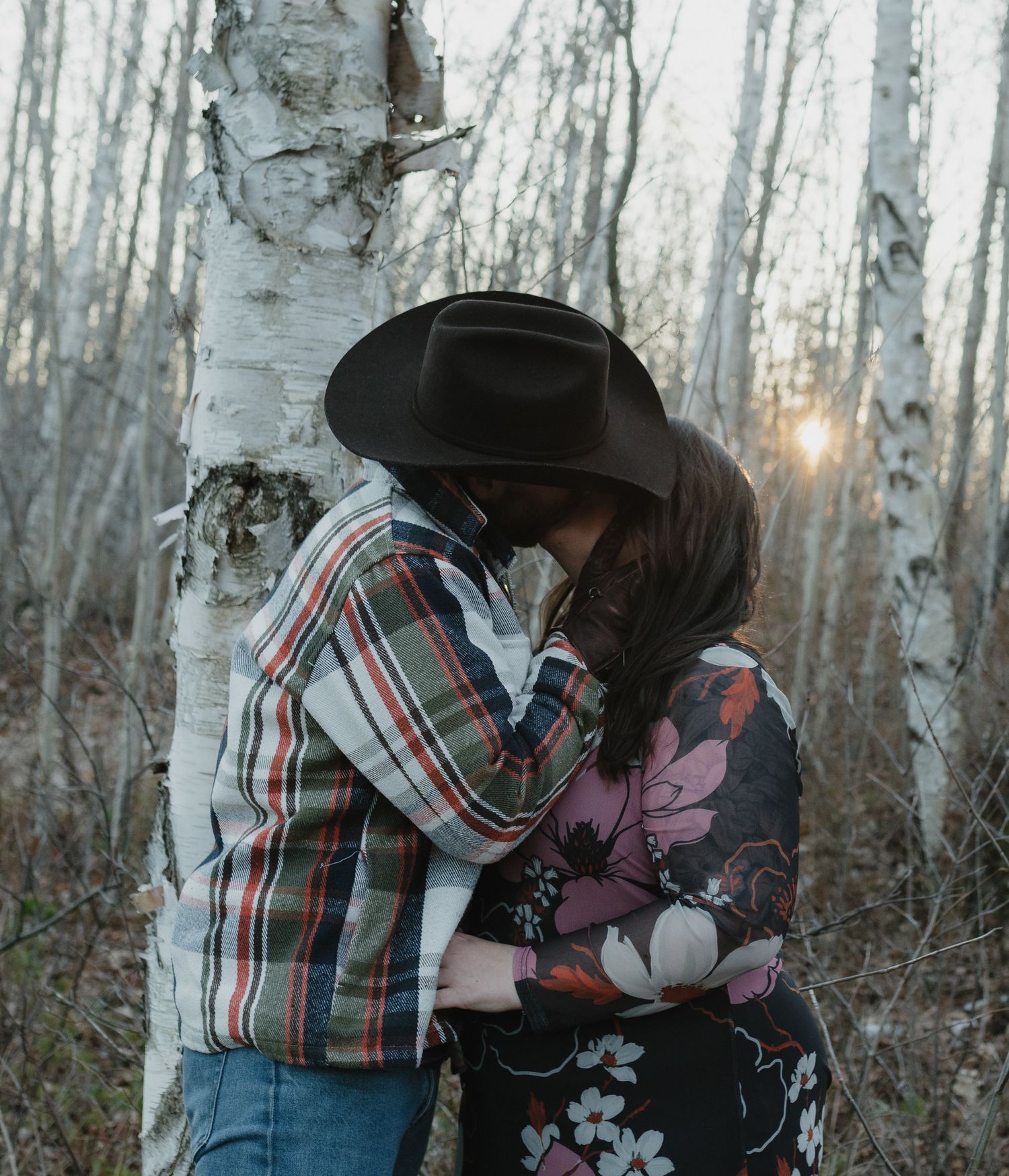 A couple in plaid and denim share a romantic moment among white birch trees in a winter forest setting.
