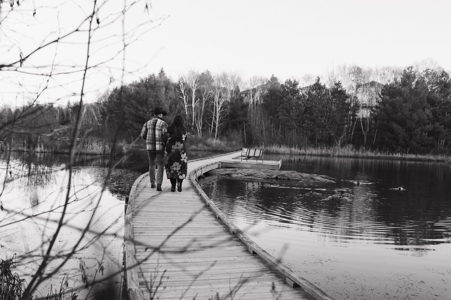 A scenic lakeside path with bare winter trees reflects in calm waters on a cloudy day.