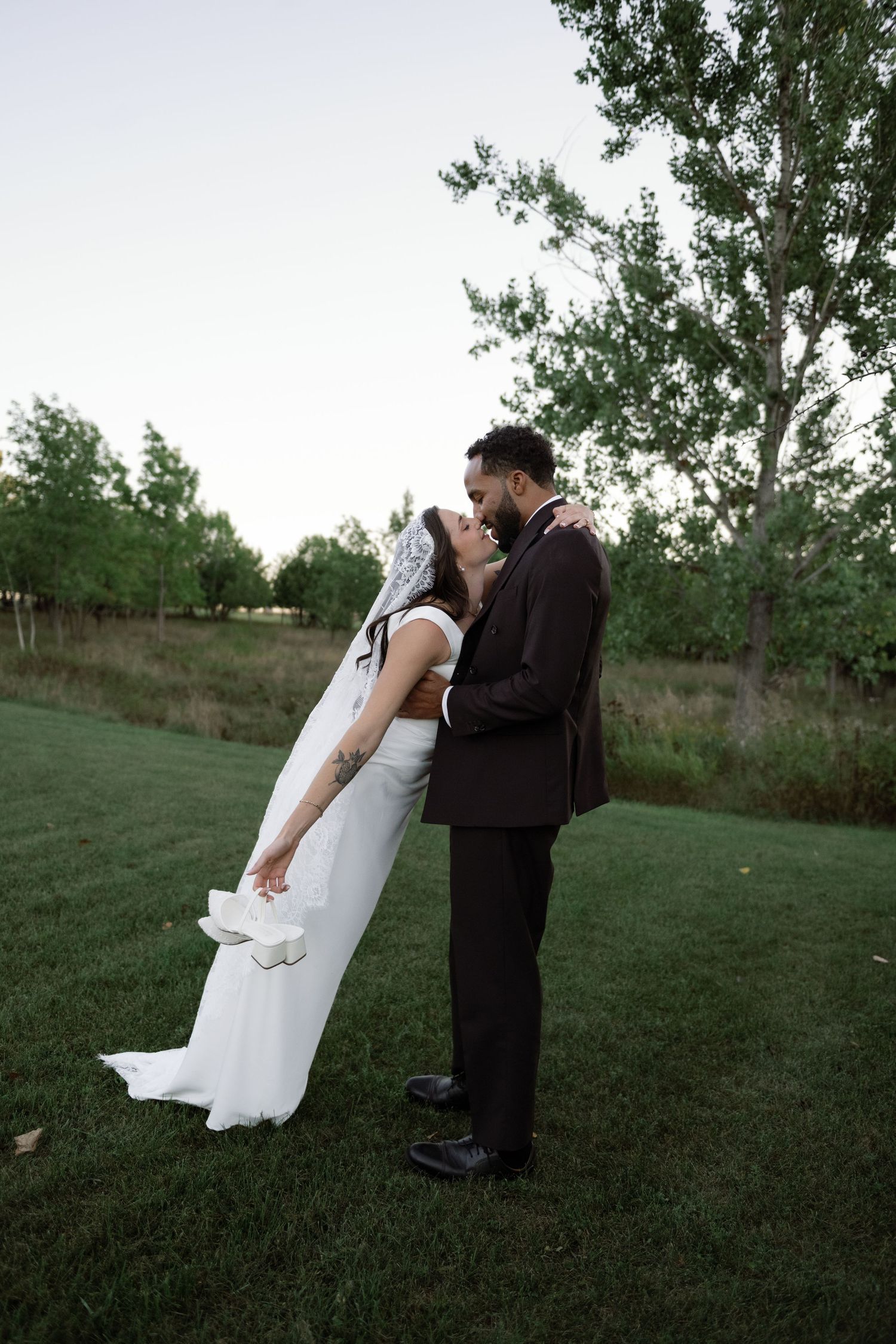 A bride and groom share a romantic kiss on a lush green lawn surrounded by trees at dusk.
