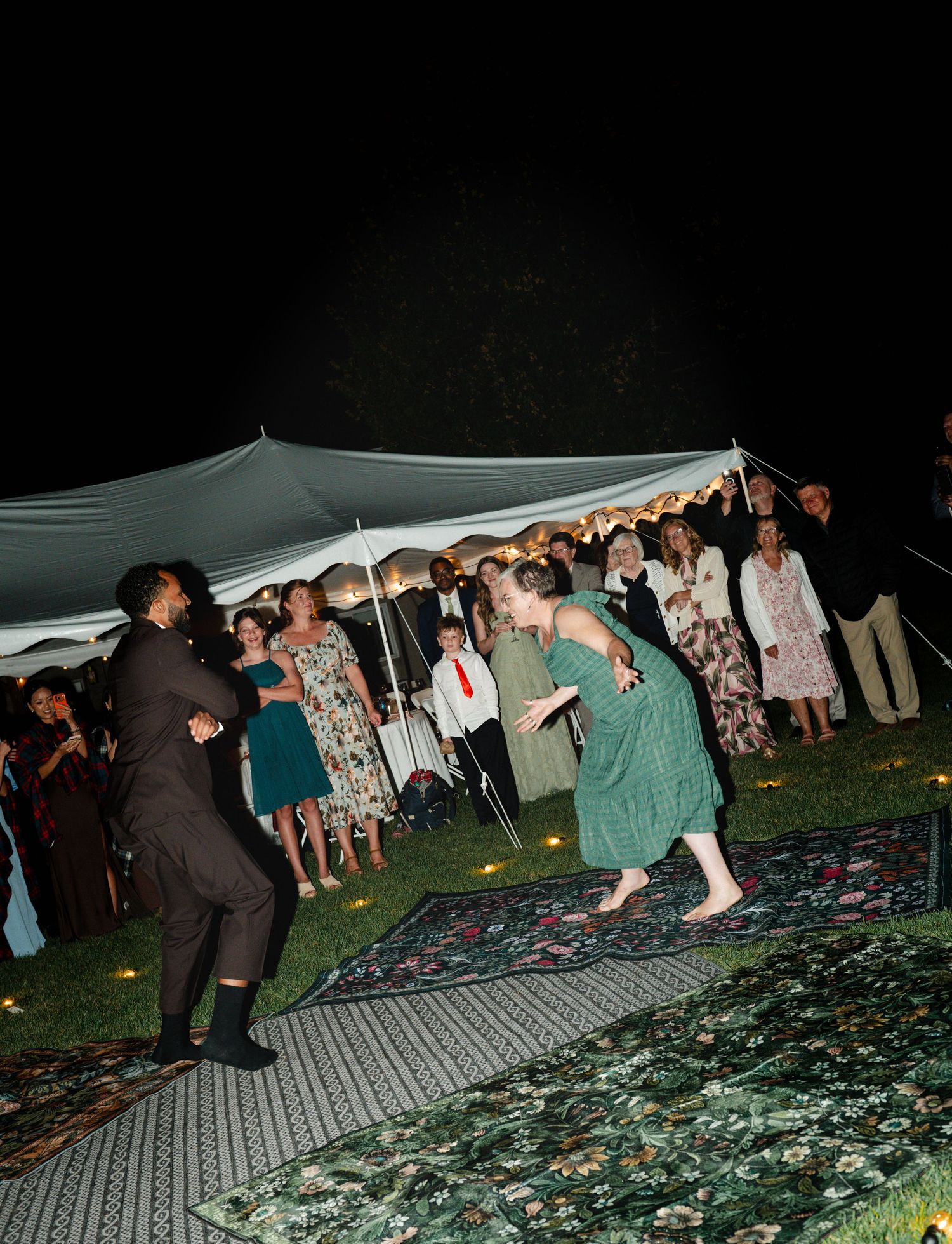 Guests dancing on a rug at an outdoor evening wedding reception under a white tent.