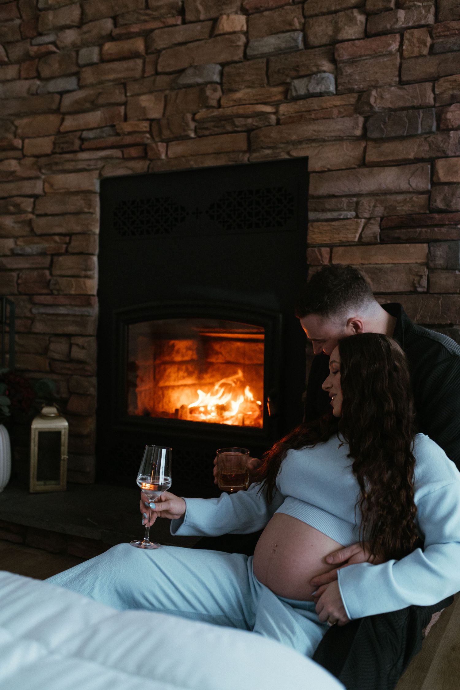 Pregnant couple relaxing by a cozy fireplace, woman holding champagne glass while partner embraces her baby bump.