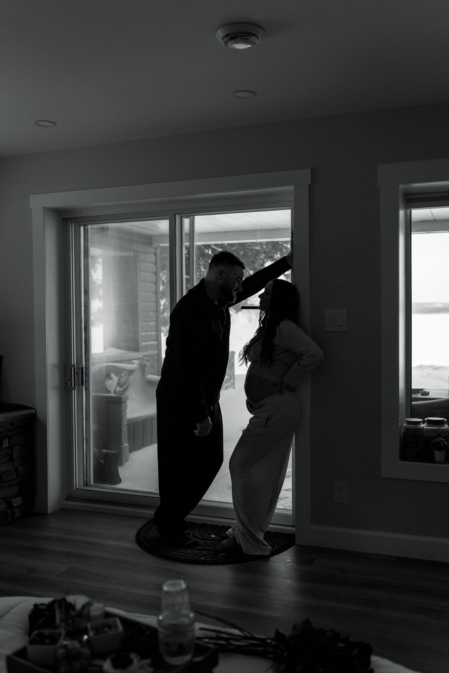 Silhouette of expecting couple sharing a kiss by sliding glass door in moody black and white maternity photo.