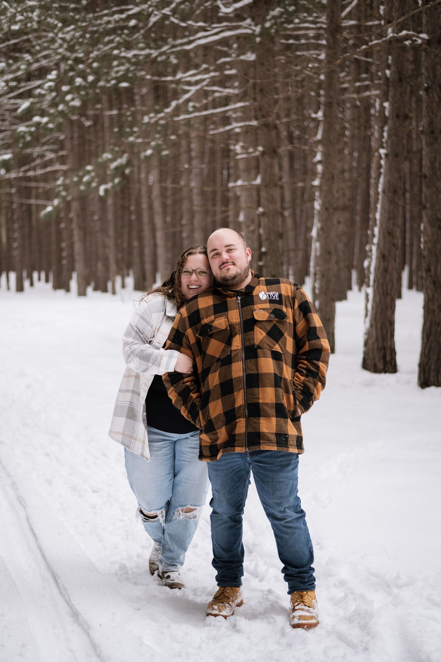 Couple smiling together in a snowy pine forest, man in plaid jacket and woman in white coat embracing.
