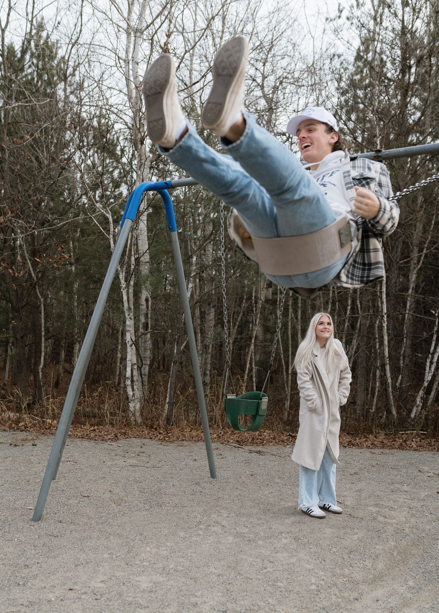 Man swinging high on playground swing while woman watches and smiles outdoors in autumn park.
