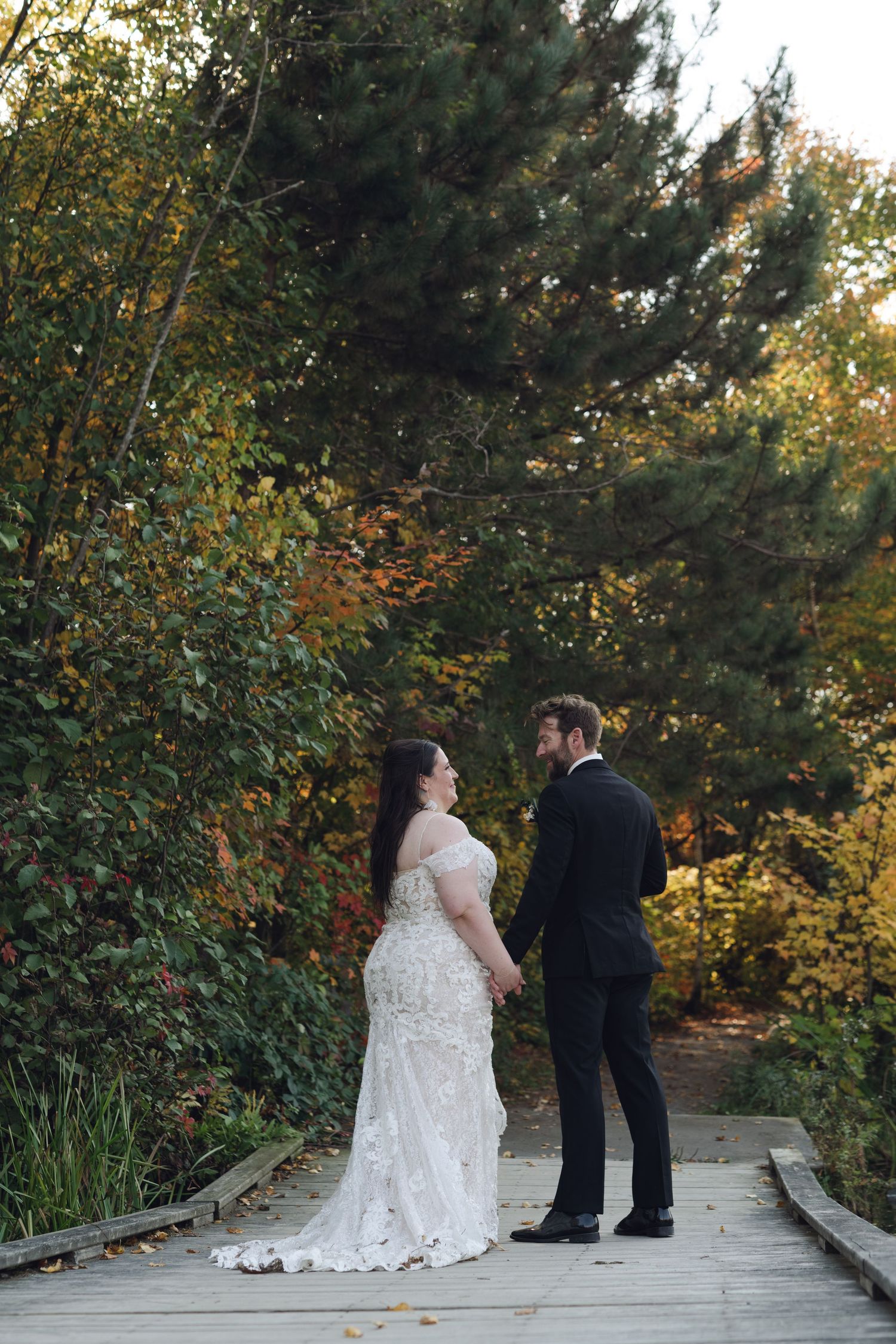 Bride and groom holding hands on a wooden bridge surrounded by autumn foliage in an outdoor garden wedding portrait.