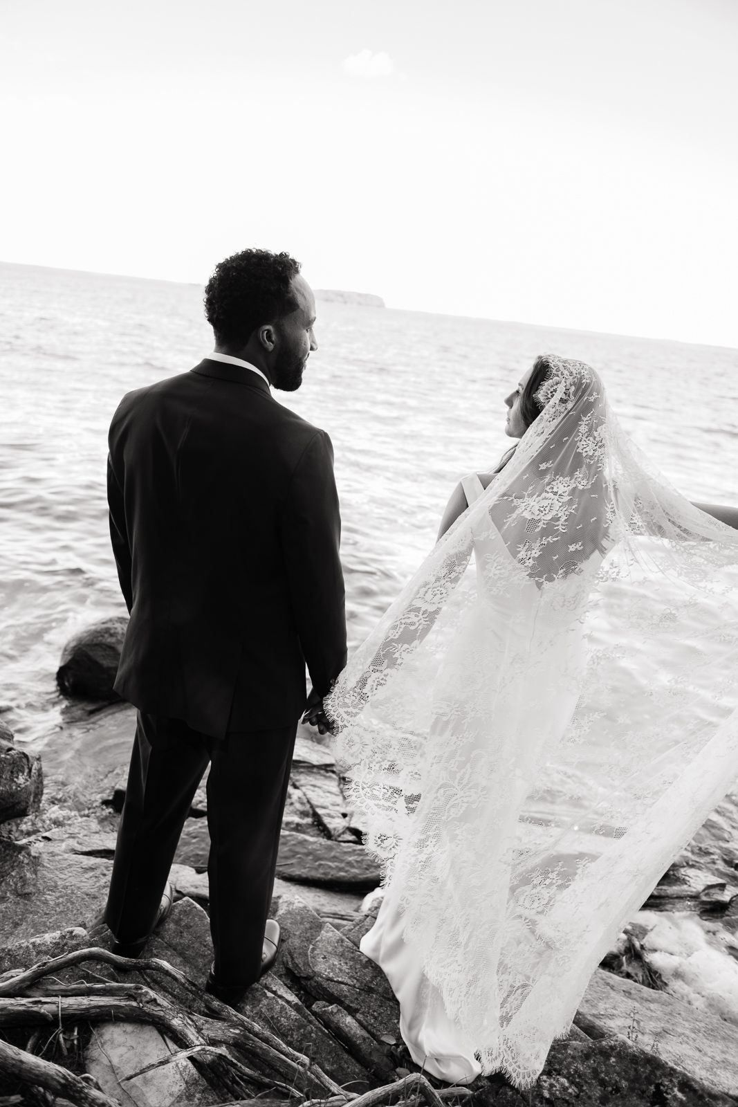 Black and white photo of a bride and groom standing on rocky shoreline, her lace veil flowing in the breeze.