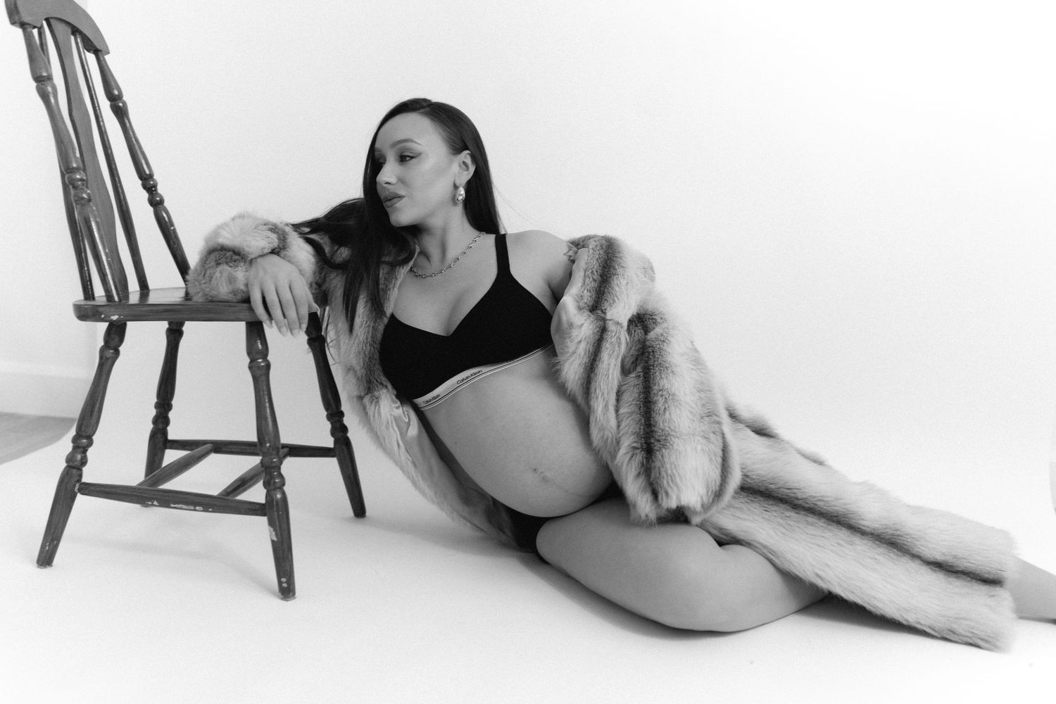 Black and white maternity photo of a pregnant woman in lingerie and fur coat posing elegantly beside a wooden chair.
