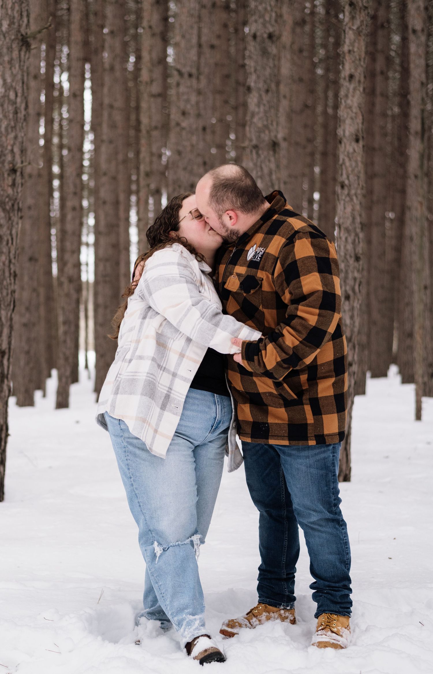 Couple kissing in snowy pine forest, wearing flannel shirts and jeans, romantic winter engagement photo.