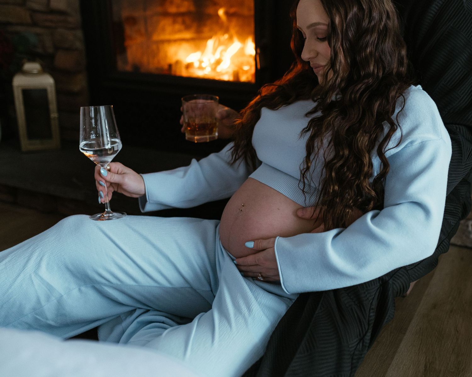 Pregnant woman in light blue outfit holding wine glass by cozy fireplace.