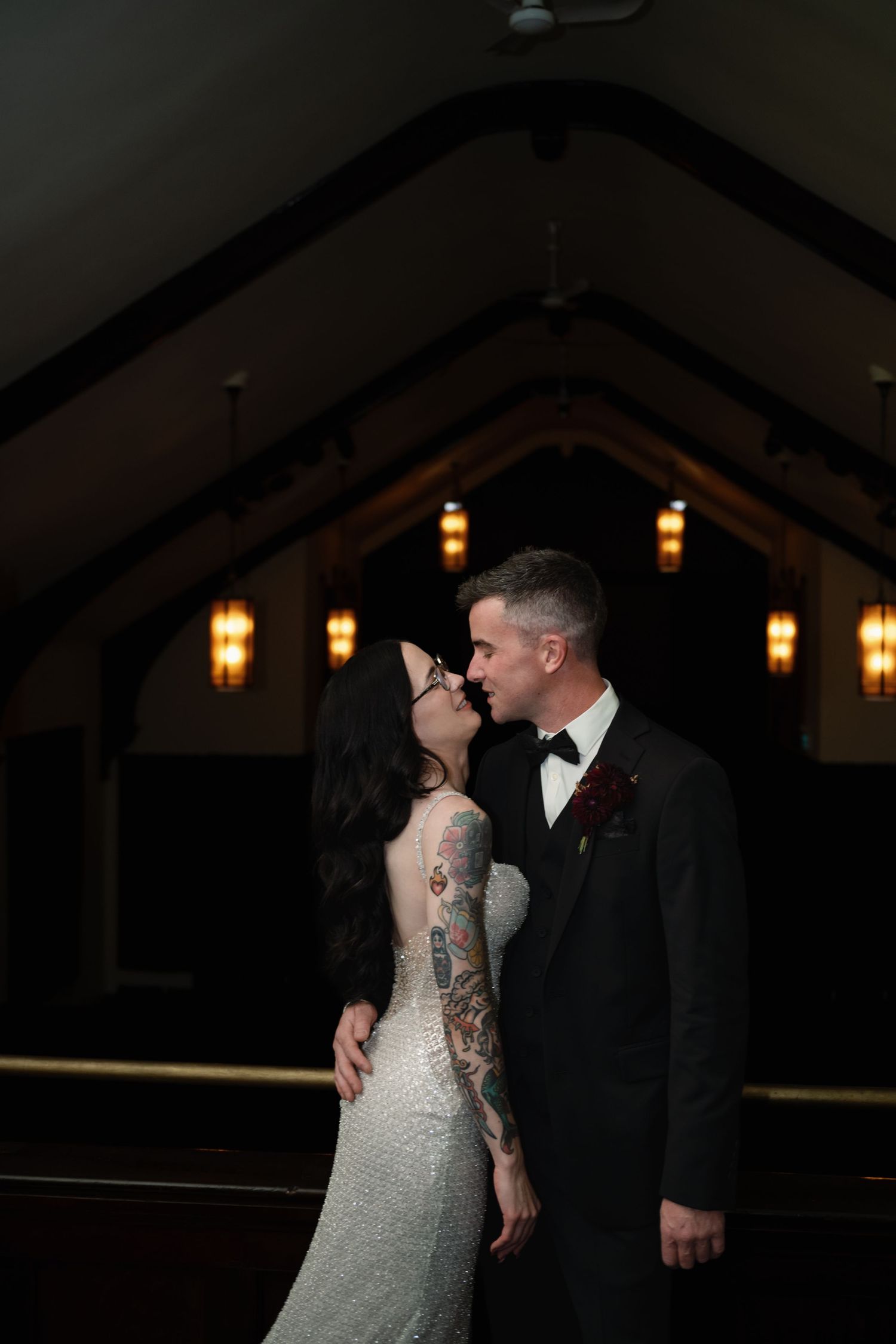 Bride with tattoos in white gown and groom in black tuxedo share a romantic kiss in a dimly lit church.