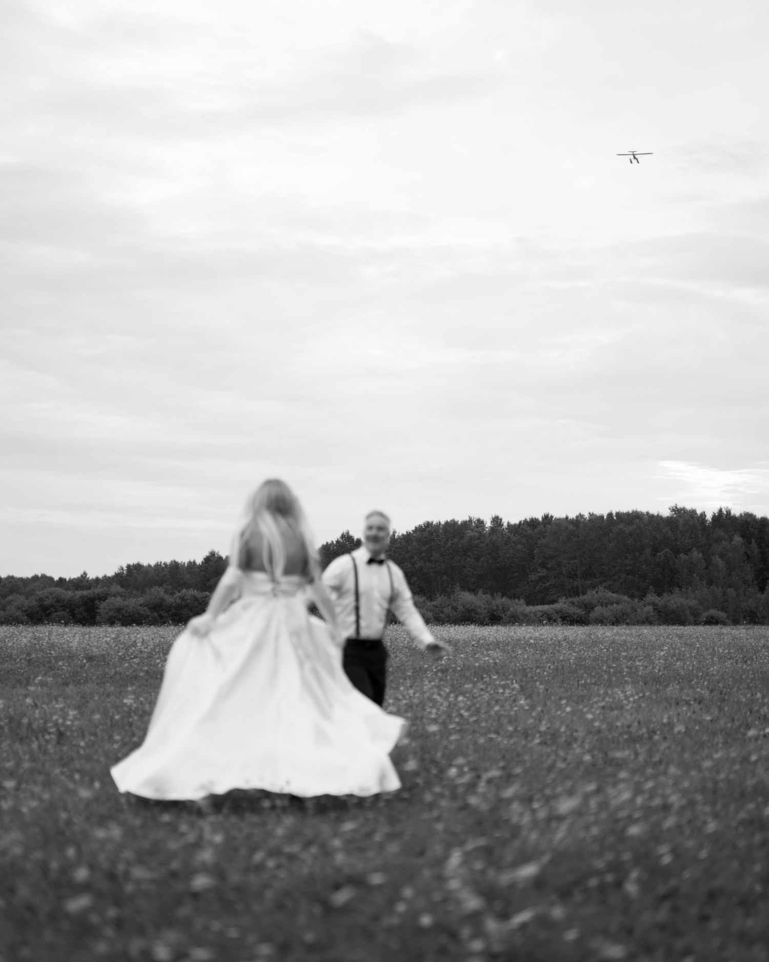 A couple in wedding attire stands in a wildflower meadow with a small airplane visible in the cloudy sky above.