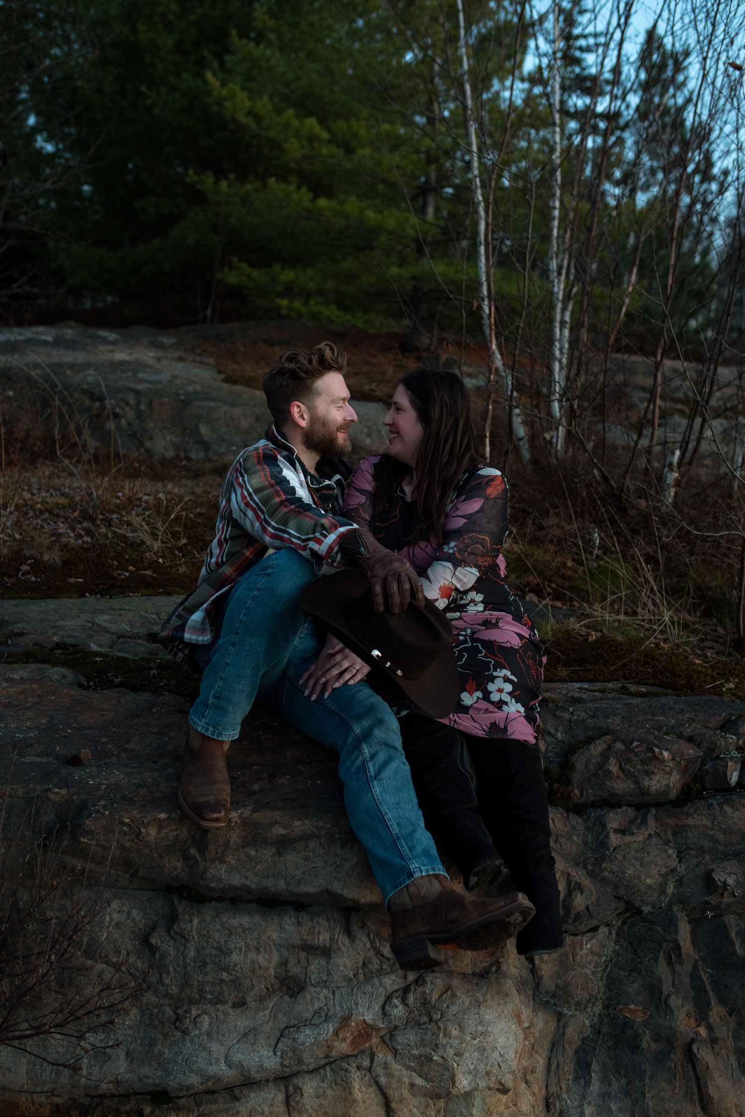 A couple sits together on a rocky cliff edge at dusk in a natural outdoor setting.