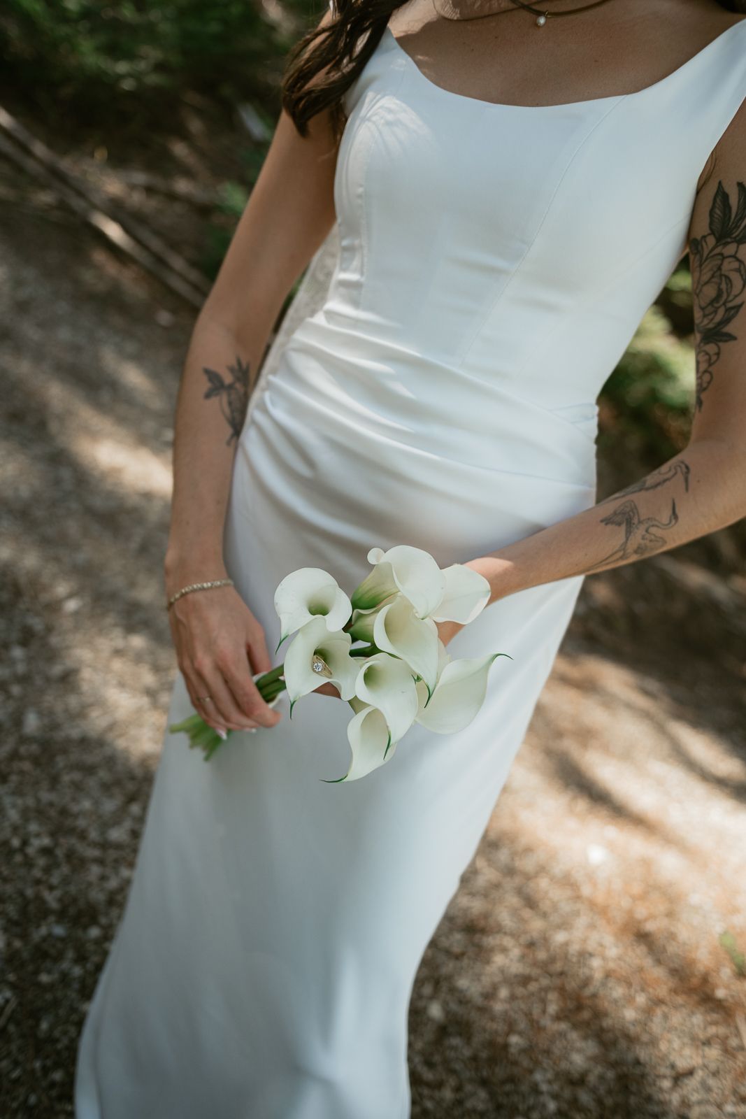 Bride in white sleeveless gown holding white calla lily bouquet, with visible tattoos on arms, standing outdoors.
