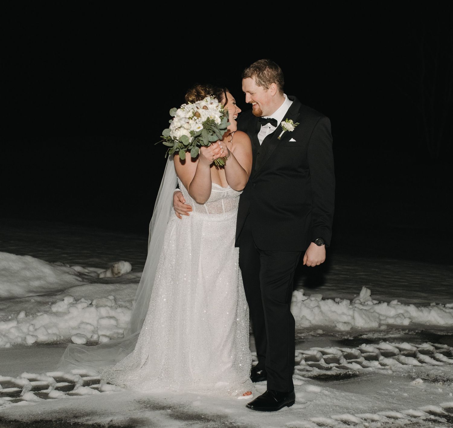 Bride and groom share a romantic moment outdoors in the snow at night, bride holding a white floral bouquet.