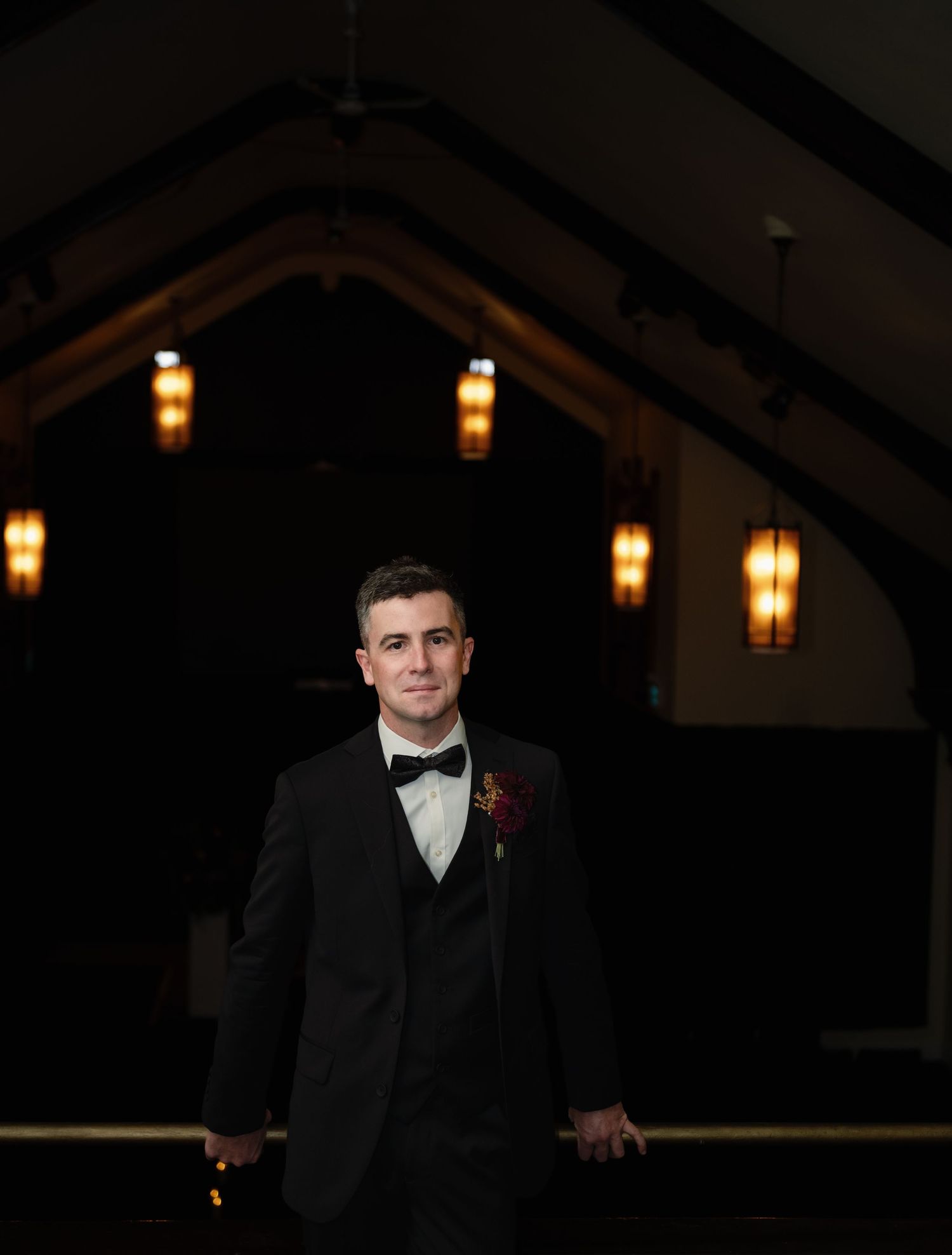 A groom in a black tuxedo with bow tie and boutonniere stands in a dimly lit wedding venue.