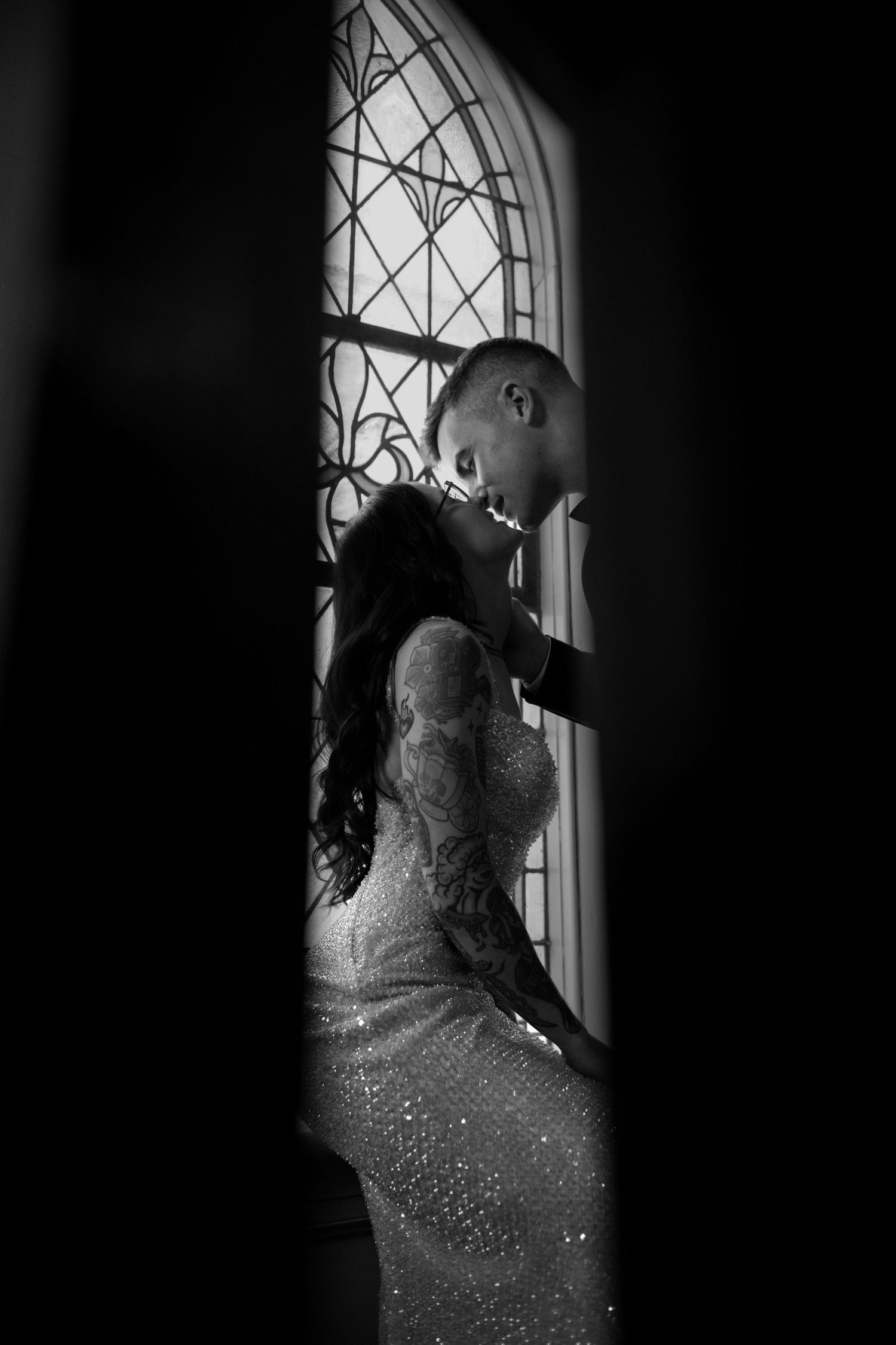 Black and white wedding photo of couple sharing intimate moment by ornate stained glass church window.