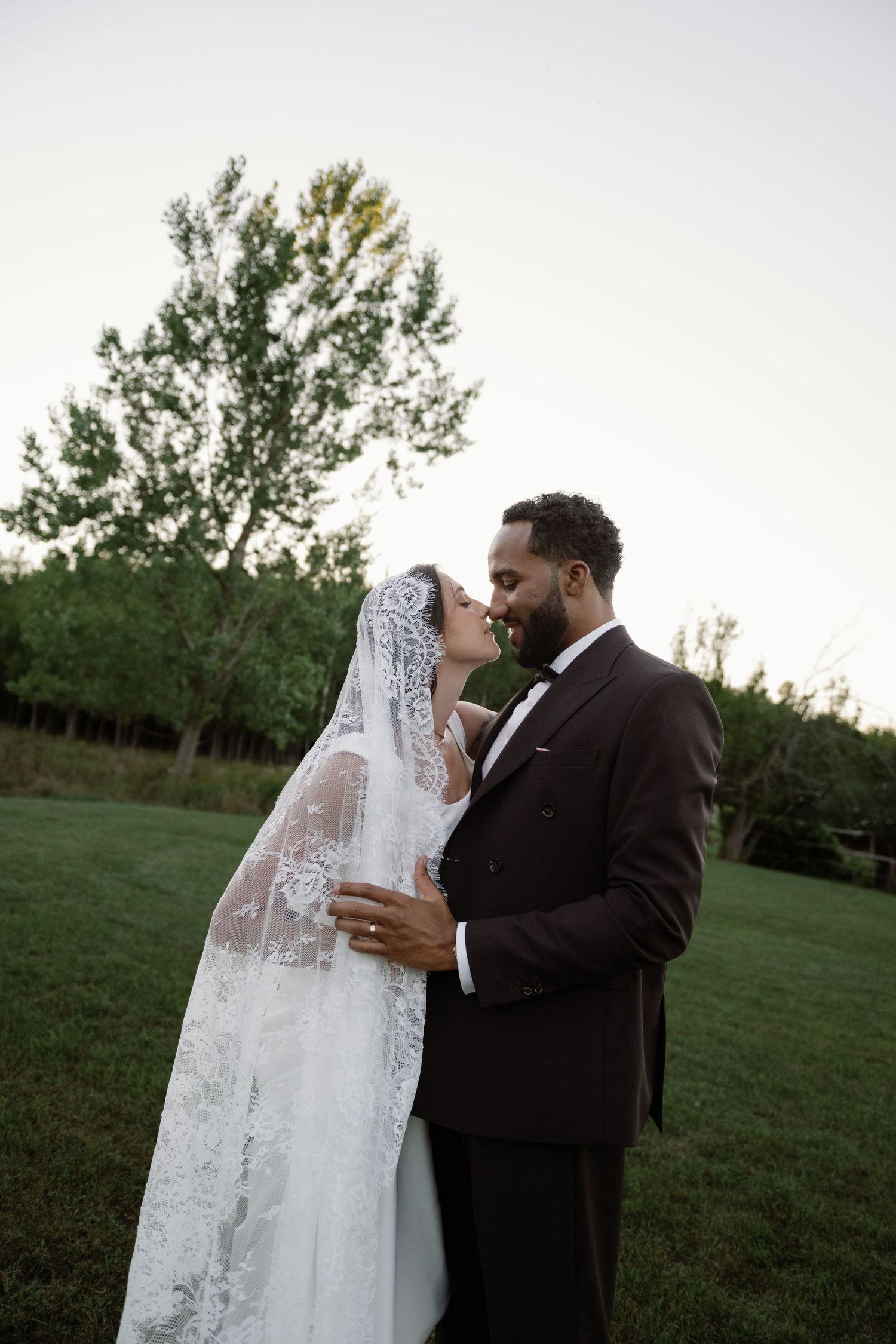 Bride and groom share a romantic kiss outdoors on green grass with trees in background at dusk.
