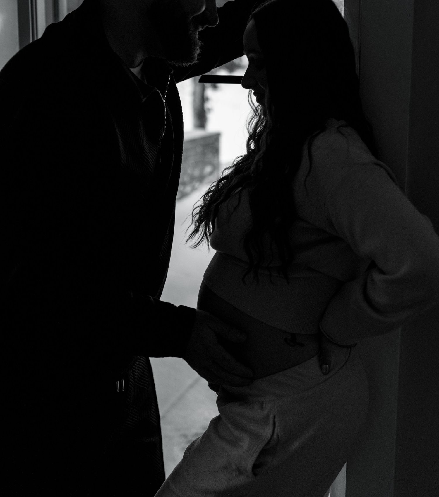 Black and white silhouette of couple embracing by window during maternity photoshoot.