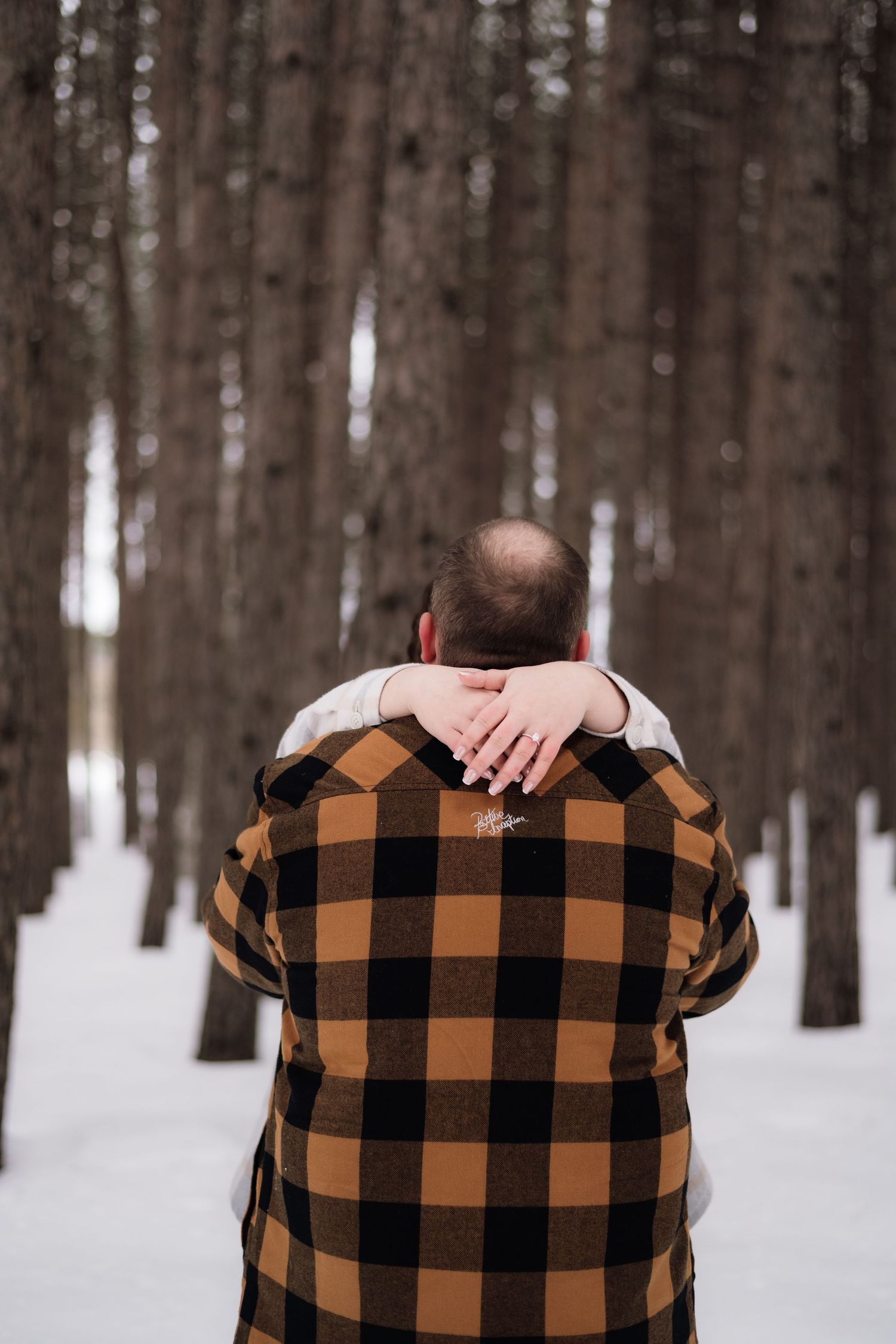 A bald man in a plaid jacket embraced from behind in a snowy pine forest, with a hand resting on his shoulder.