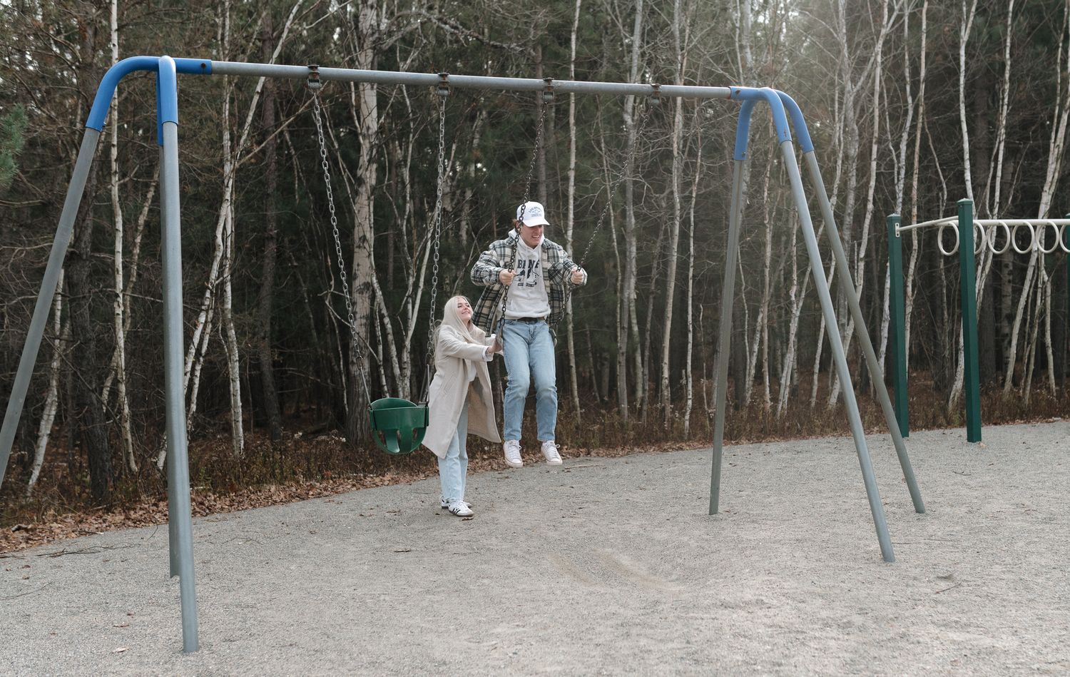Two people at an outdoor playground swing set surrounded by bare autumn trees.
