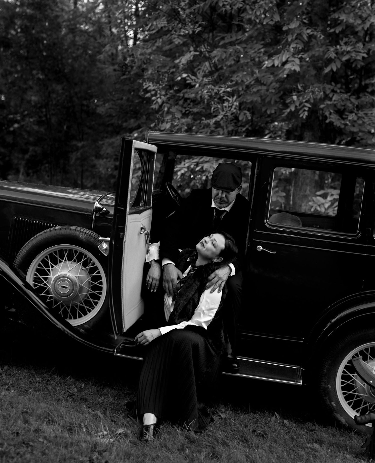 Vintage black and white photo of couple posing dramatically by a classic 1930s automobile in a wooded setting.